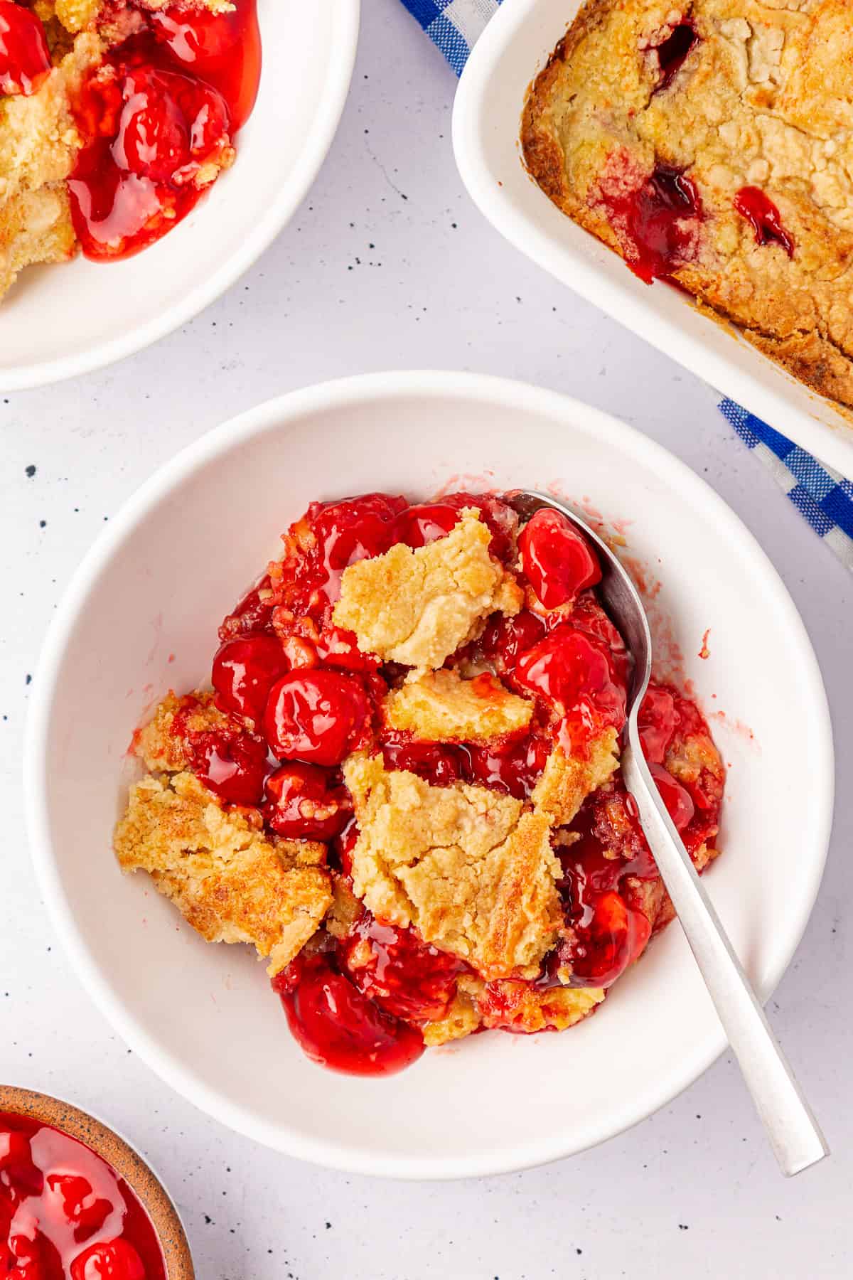 A bowl of cherry dump cake cobbler with a golden, crumbly crust is shown. A spoon rests in the bowl, surrounded by part of a baking dish and another bowl filled with tempting cobbler on a white speckled surface.