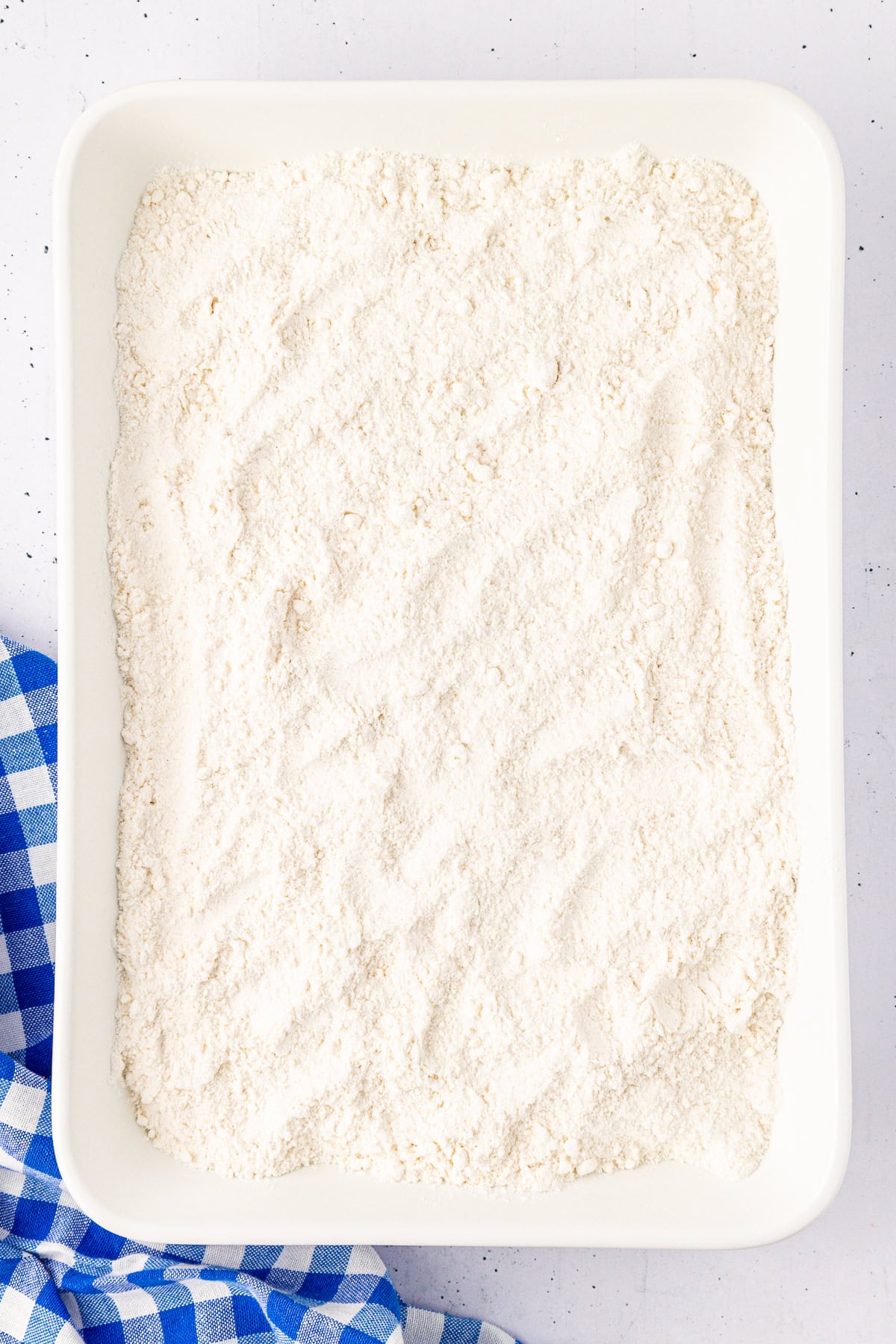 A rectangular white tray, perhaps poised for a cherry dump cake creation, is filled with a fine, white powdery substance—likely flour. It rests on a speckled surface, with the corner graced by a blue and white checkered cloth peeking in.