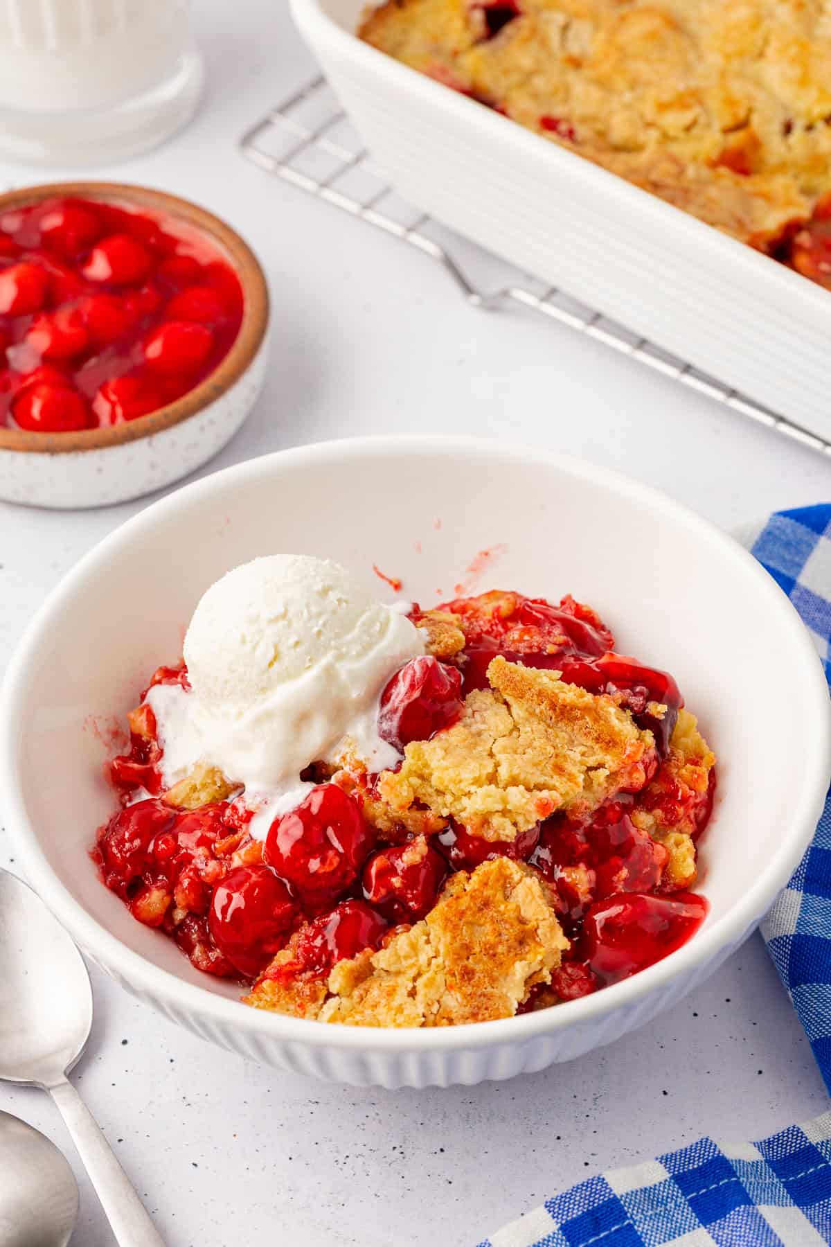 A bowl of cherry cobbler topped with a scoop of vanilla ice cream. The dish features golden, crumbly crust pieces and bright red cherries reminiscent of a cherry dump cake. In the background, a dish of cherry filling and a baking dish with cobbler are visible against a blue-checkered cloth.