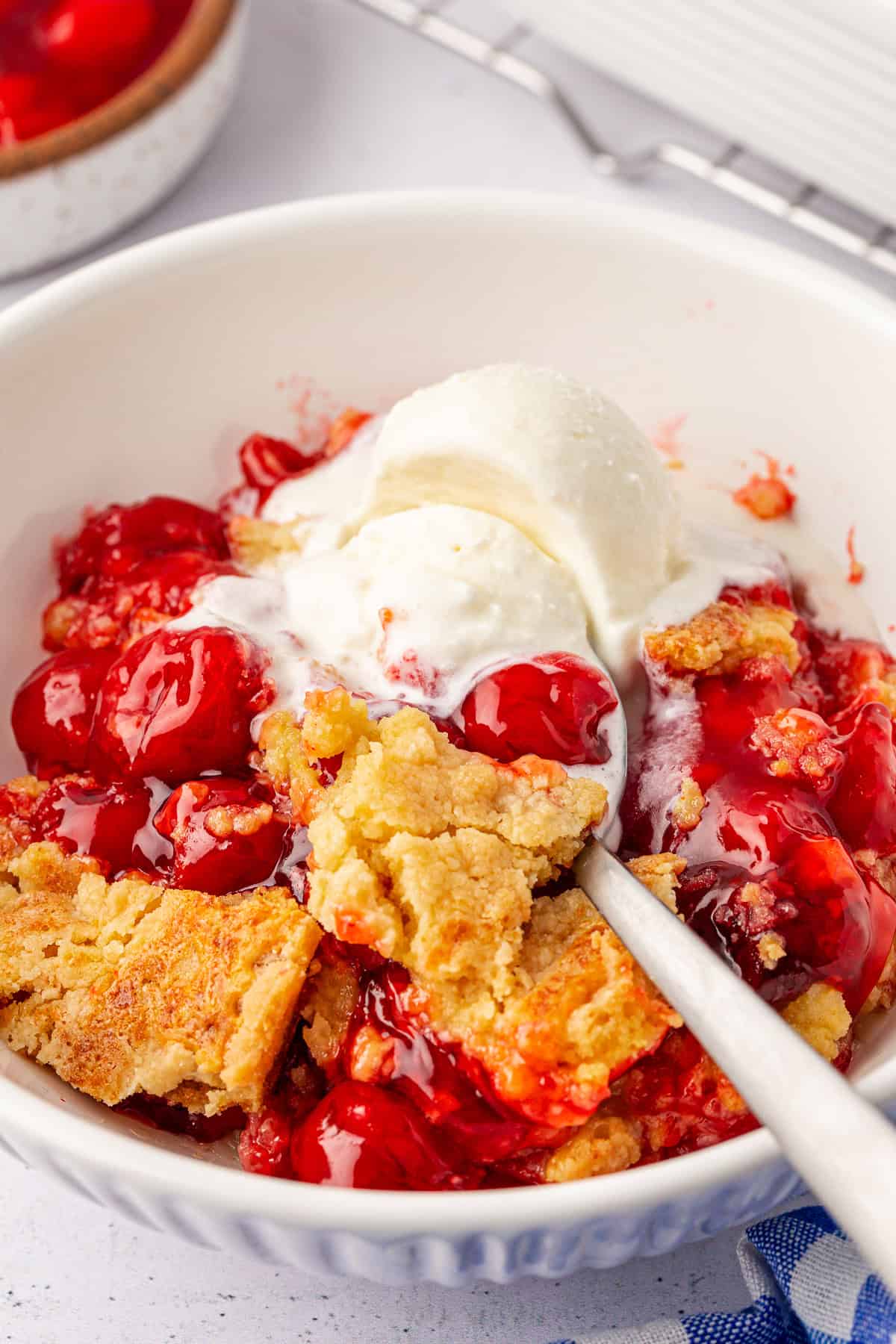 A close-up of a bowl filled with cherry cobbler and a scoop of vanilla ice cream on top. The cobbler, reminiscent of a cherry dump cake, boasts a golden-brown crust with vibrant red cherries, and a spoon rests in the dish. A hint of a blue cloth is visible at the bottom corner.