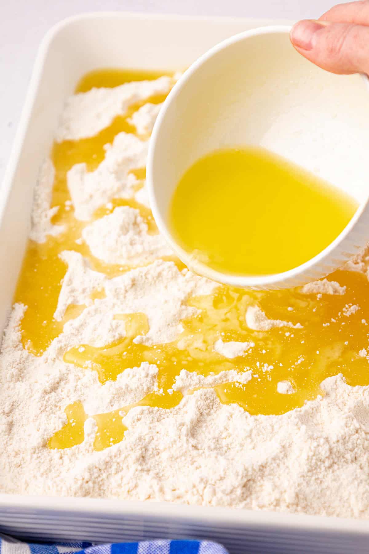 A hand pours melted butter from a white cup over a mix of flour in a rectangular white dish, setting the stage for a cherry dump cake delight. A blue and white striped cloth peeks out beneath the dish.