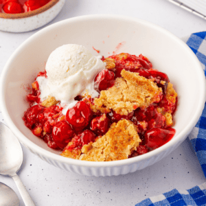 A bowl of cherry cobbler crowned with a scoop of vanilla ice cream resembles a cherry dump cake. The dessert is vibrant red, punctuated by chunks of golden-brown crust. Nearby, a blue-checked napkin and two silver spoons rest on the pristine white surface.