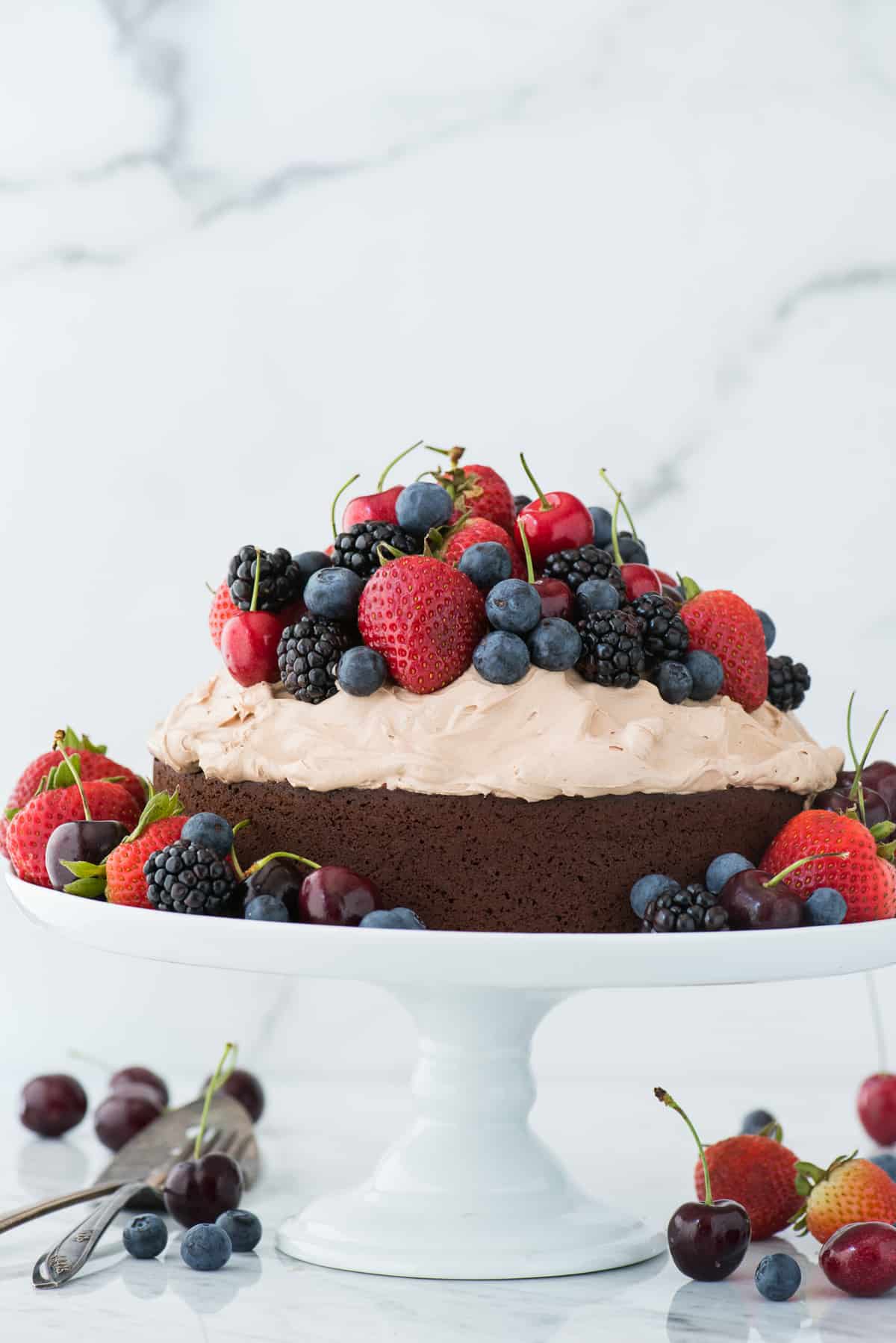 chocolate cake with whipped frosting topped with fresh cherries, strawberries, and blackberries on white cake stand on white background