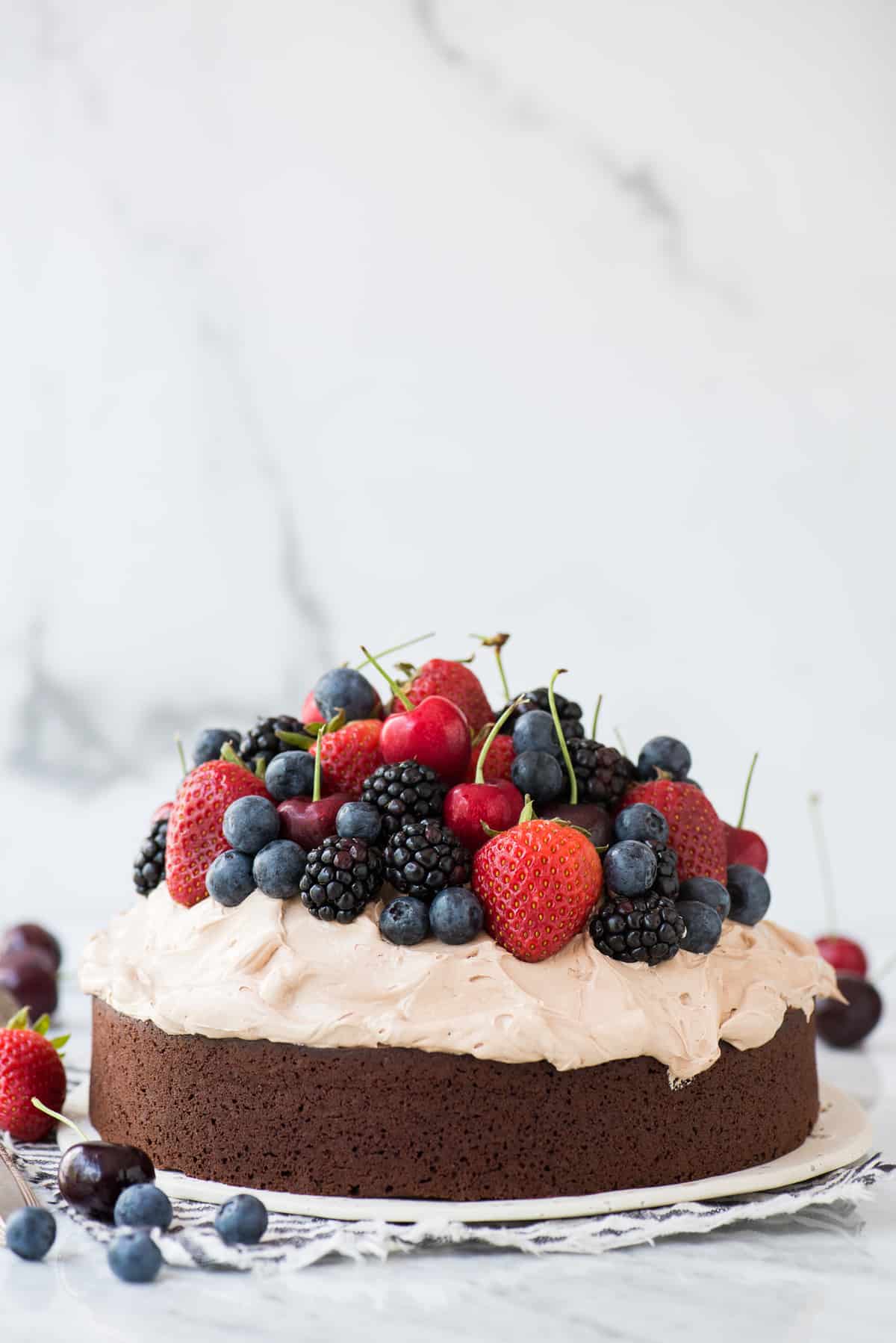 chocolate cake with whipped frosting topped with fresh cherries, strawberries, and blackberries on white plate on white background