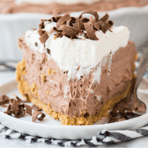 A slice of chocolate pudding pie with a graham cracker crust, creamy chocolate filling, whipped cream topping, and chocolate shavings, served on a white plate with a fork.