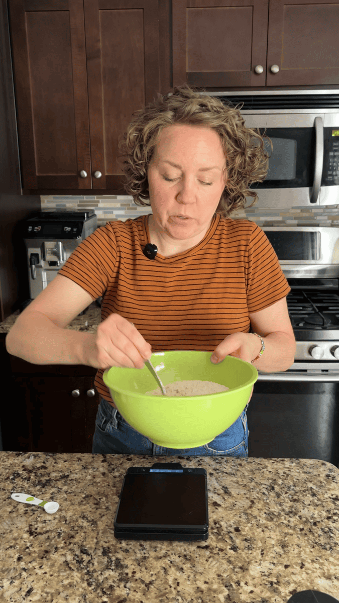 A woman in a striped shirt stirs ingredients for pumpkin sugar cookies in a green bowl, standing in a kitchen with dark cabinets and stainless steel appliances.