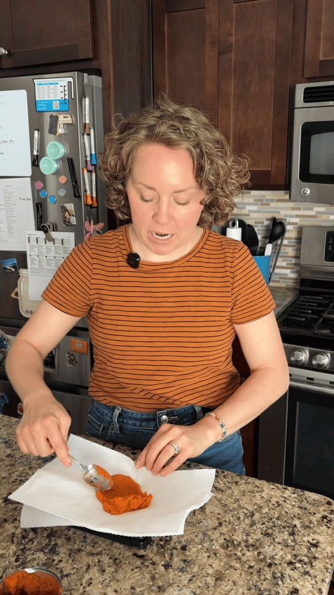 A person with curly hair in a striped shirt mashes orange food—perhaps the filling for pumpkin sugar cookies—with a fork on a plate in a kitchen with wooden cabinets and stainless steel appliances.