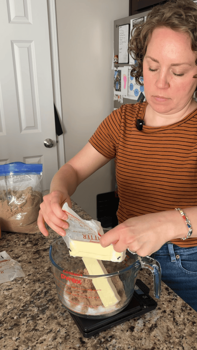 A woman in a striped shirt puts butter into a glass measuring cup on a kitchen counter, preparing to bake pumpkin sugar cookies, with a bag of brown sugar and other ingredients nearby.
