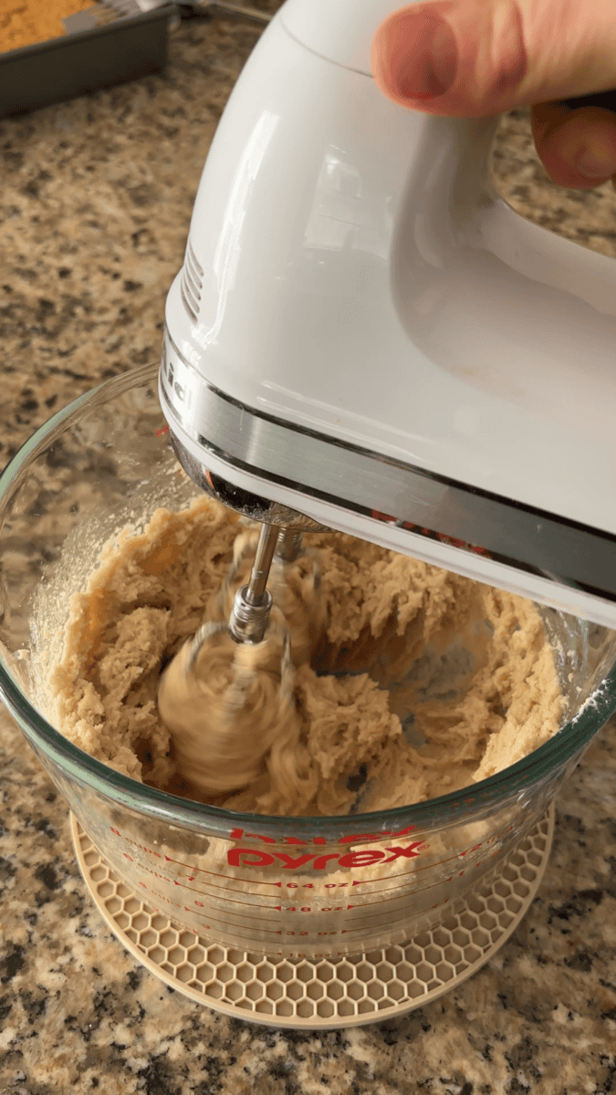 A hand holds a white electric mixer blending pumpkin sugar cookie dough in a glass Pyrex measuring bowl on a granite countertop.