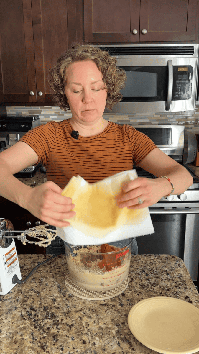 A person in a striped shirt holds a yellow-stained paper towel over a mixing bowl in a kitchen, surrounded by baking ingredients and utensils on the counter—perfectly set up for making pumpkin sugar cookies.