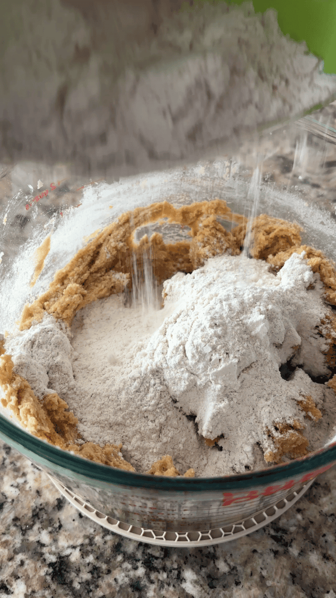 Flour is being poured into a glass bowl containing a creamed mixture, likely for baking pumpkin sugar cookies or a cake, on a speckled countertop.