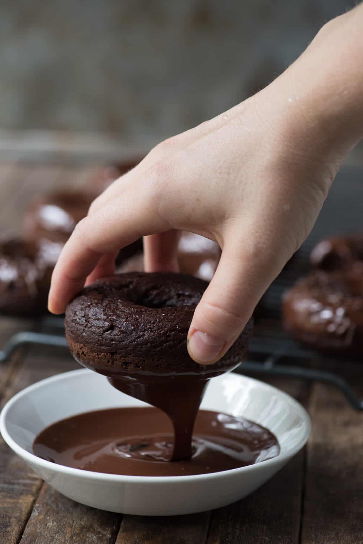 chocolate donut being dipped into chocolate glaze in white bowl