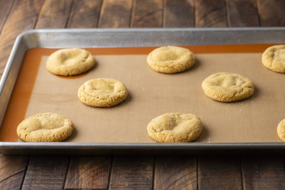 A baking sheet with eight golden brown pumpkin sugar cookies resting on a silicone baking mat, set on a wooden surface.