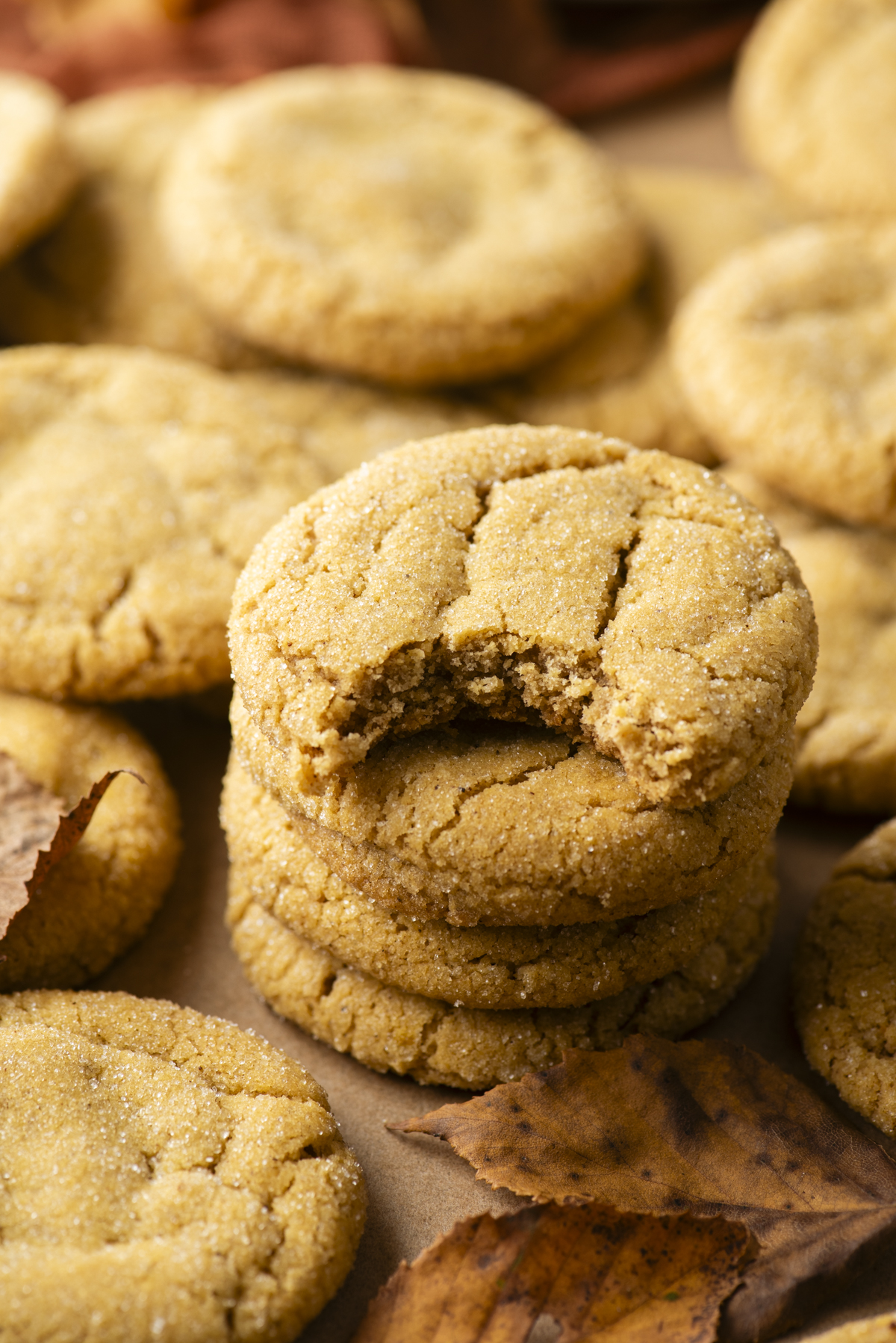 A stack of three pumpkin sugar cookies, the top one with a bite taken out of it, surrounded by more cookies and autumn leaves.