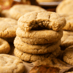 A stack of three pumpkin sugar cookies sits on a table, with the top cookie missing a bite. More cookies and autumn leaves surround the stack.