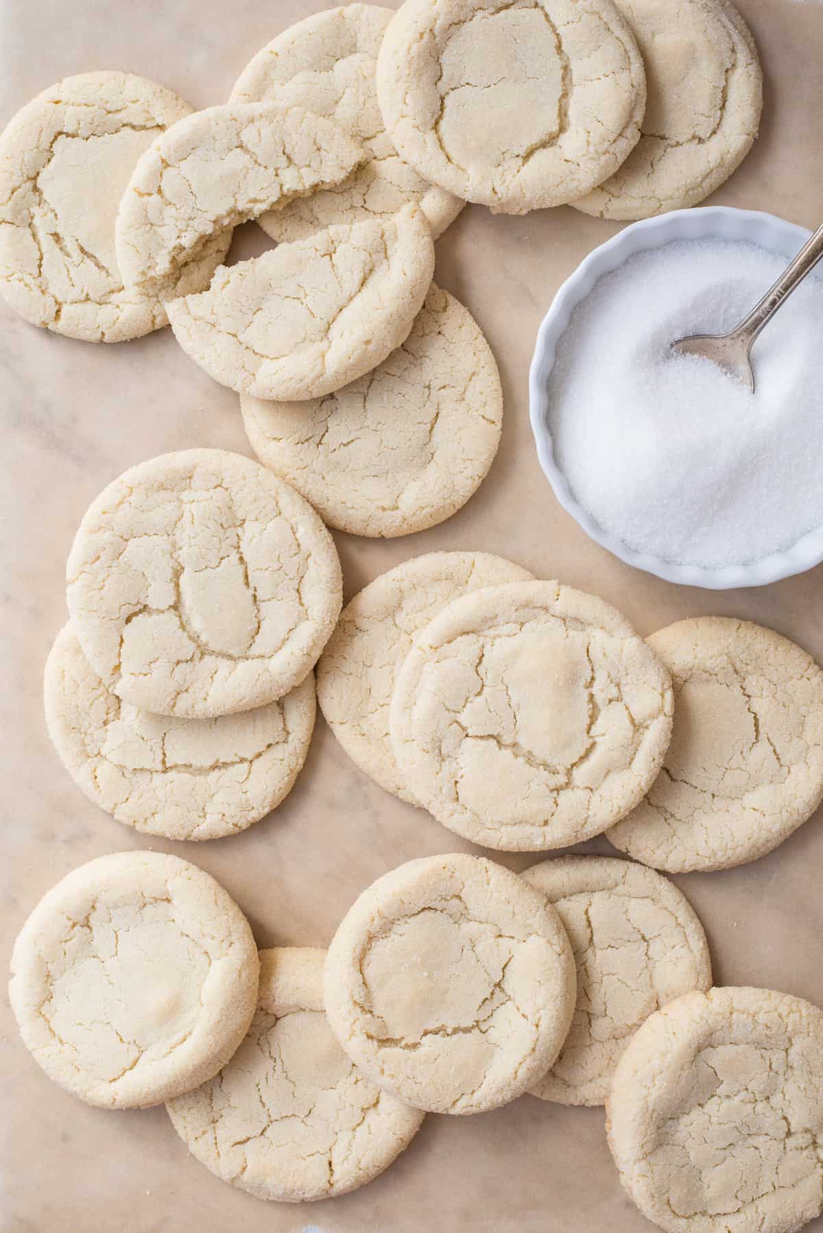 drop sugar cookies scattered on brown parchment paper with bowl of sugar on right side