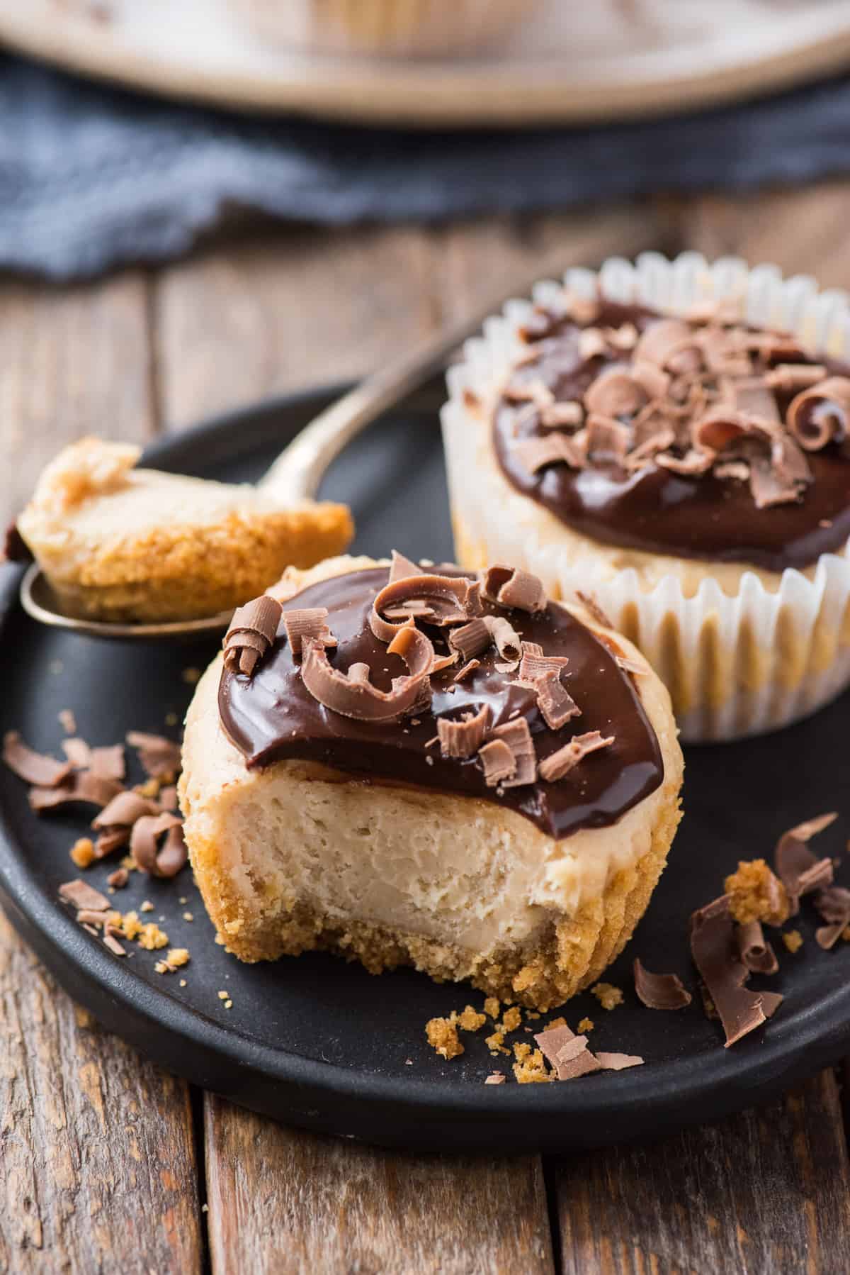 two mini peanut butter cheesecakes with chocolate ganache topping dark plate on wood background