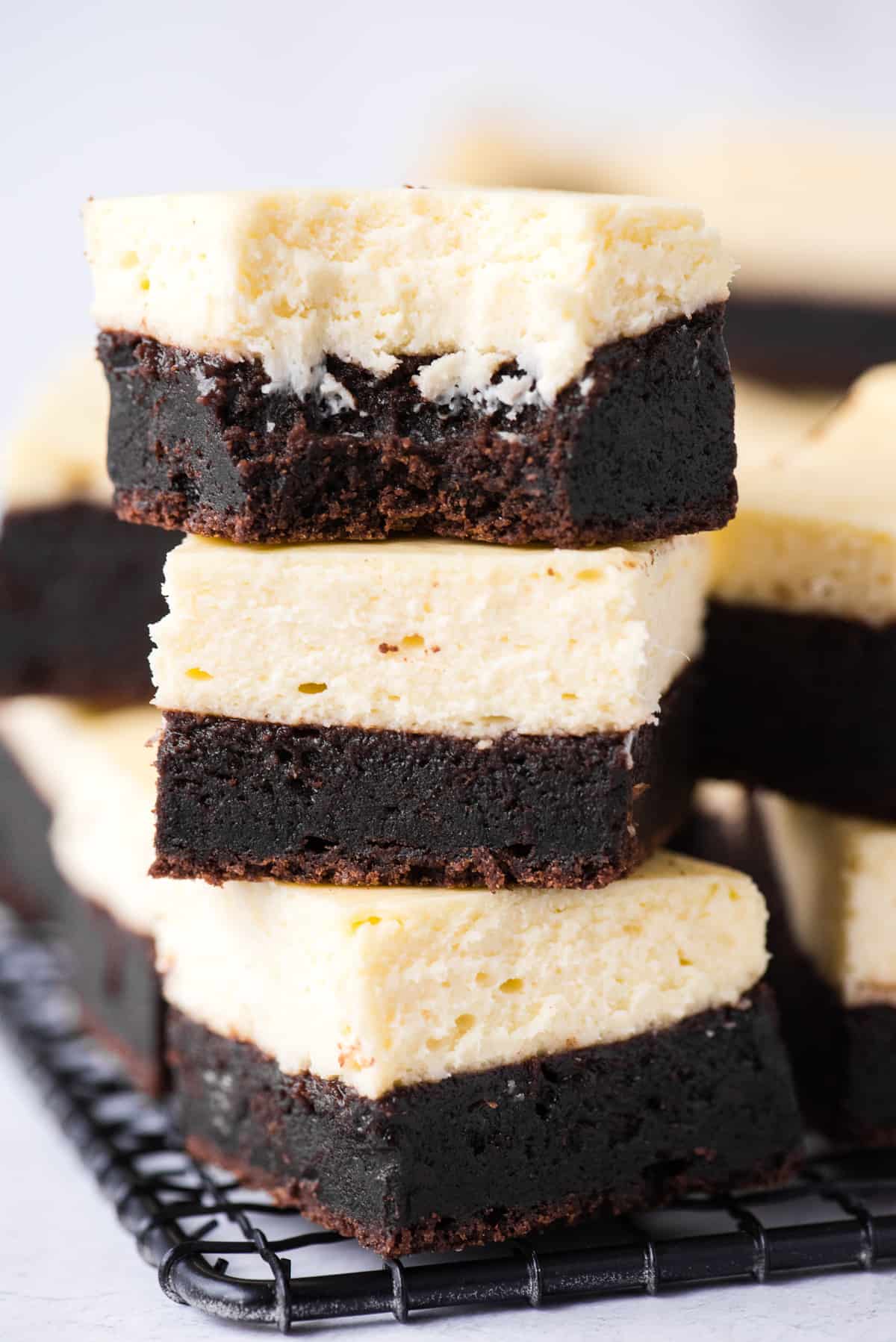 three cheesecake brownies stacked on each other, top brownie has bite removed on black wire rack on white background