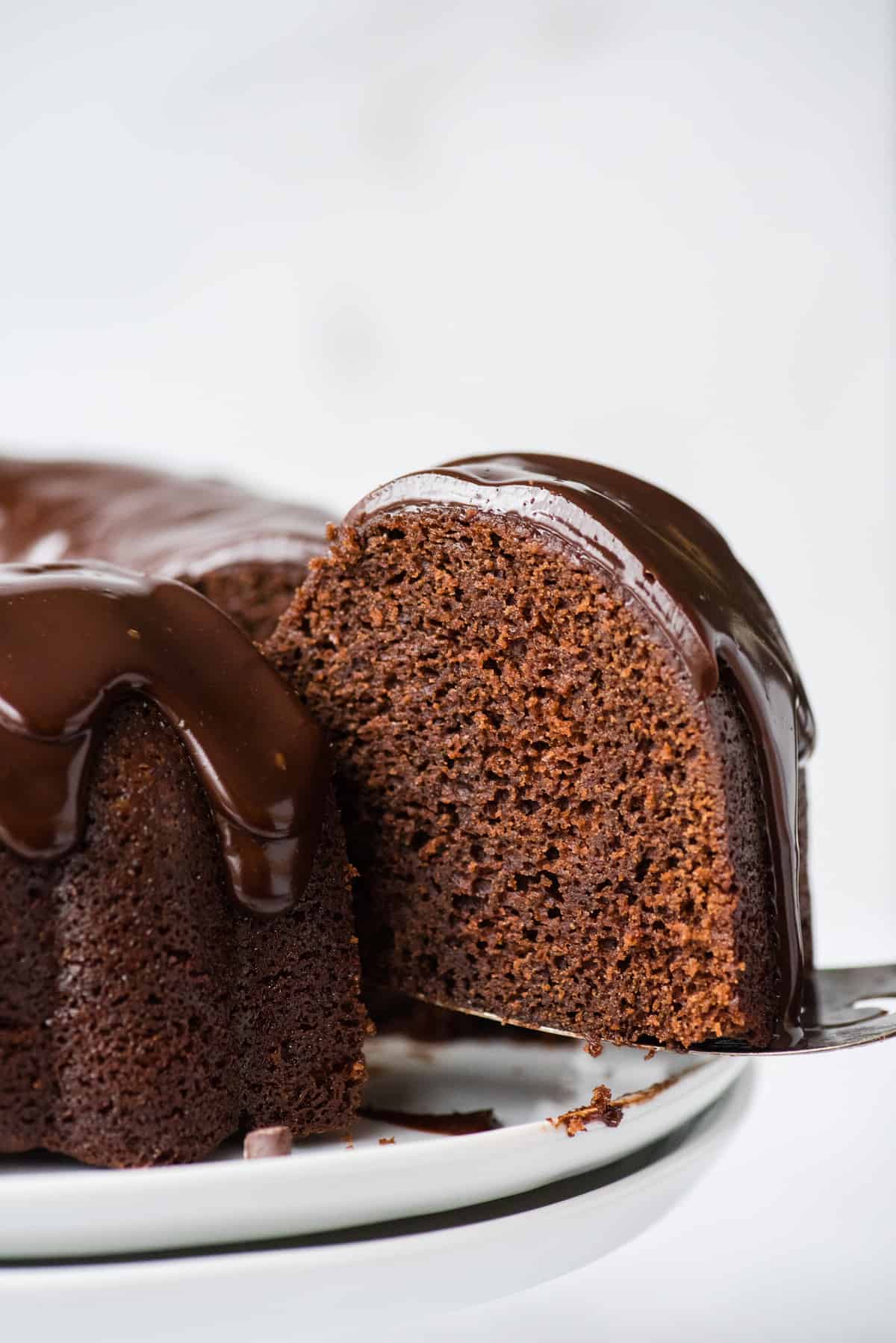 slice of chocolate bundt cake with chocolate glaze being removed from the cake on metal spatula on white background