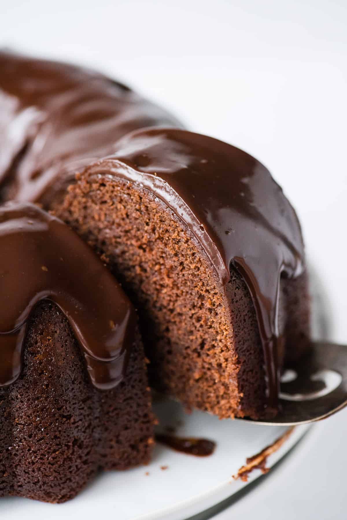slice of chocolate bundt cake with chocolate glaze being removed from the cake on metal spatula on white background