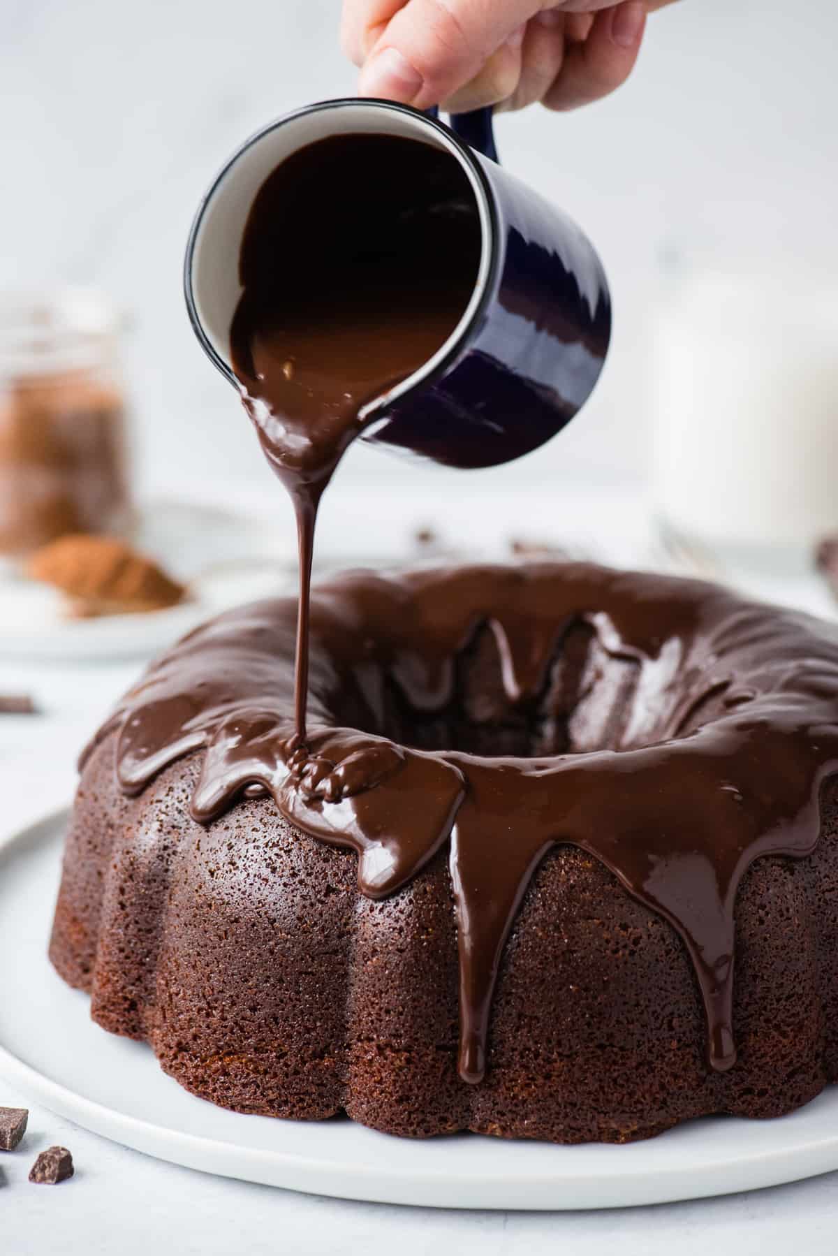 chocolate glaze being poured over chocolate bundt cake on white plate on white background