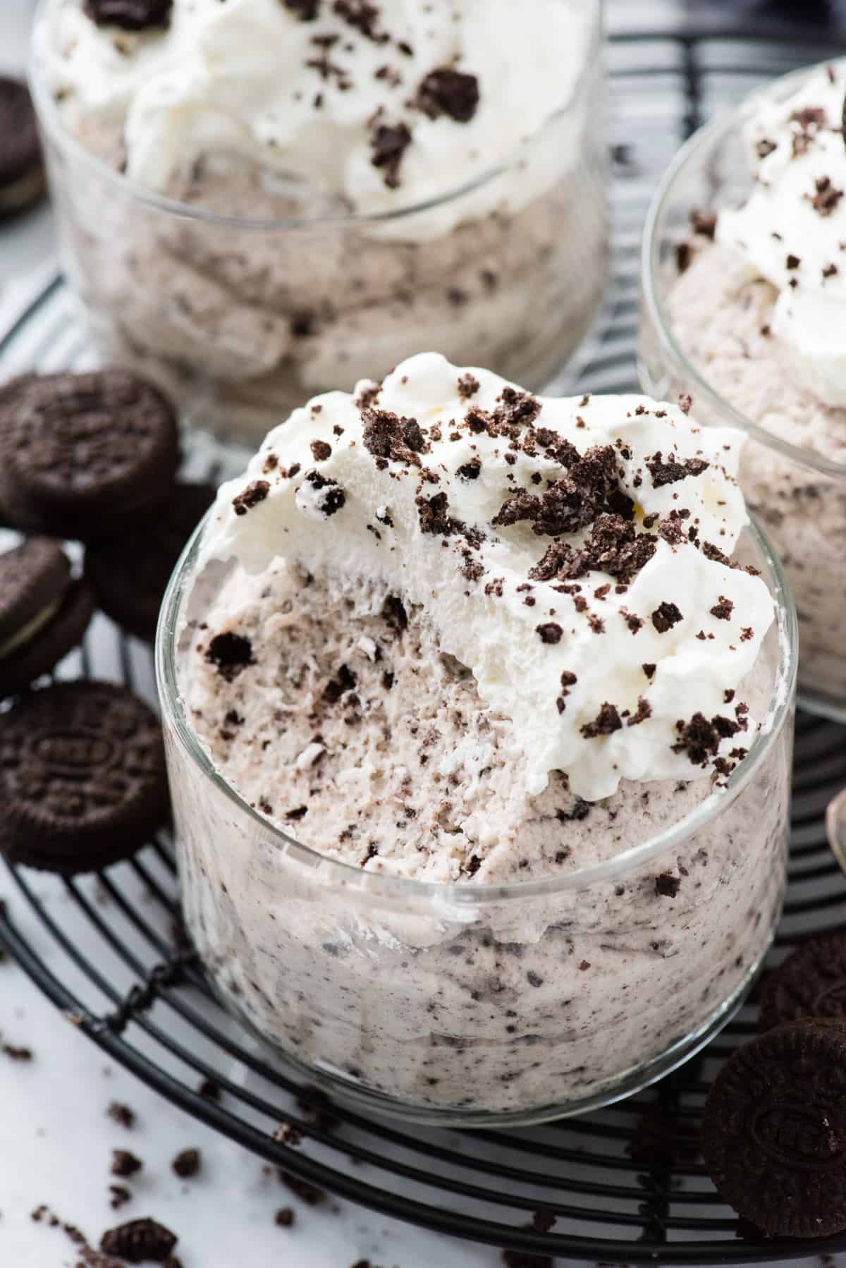 oreo mousse with scoop removed in glass dessert bowl topped with whipped cream and oreo pieces on black circle rack on white background