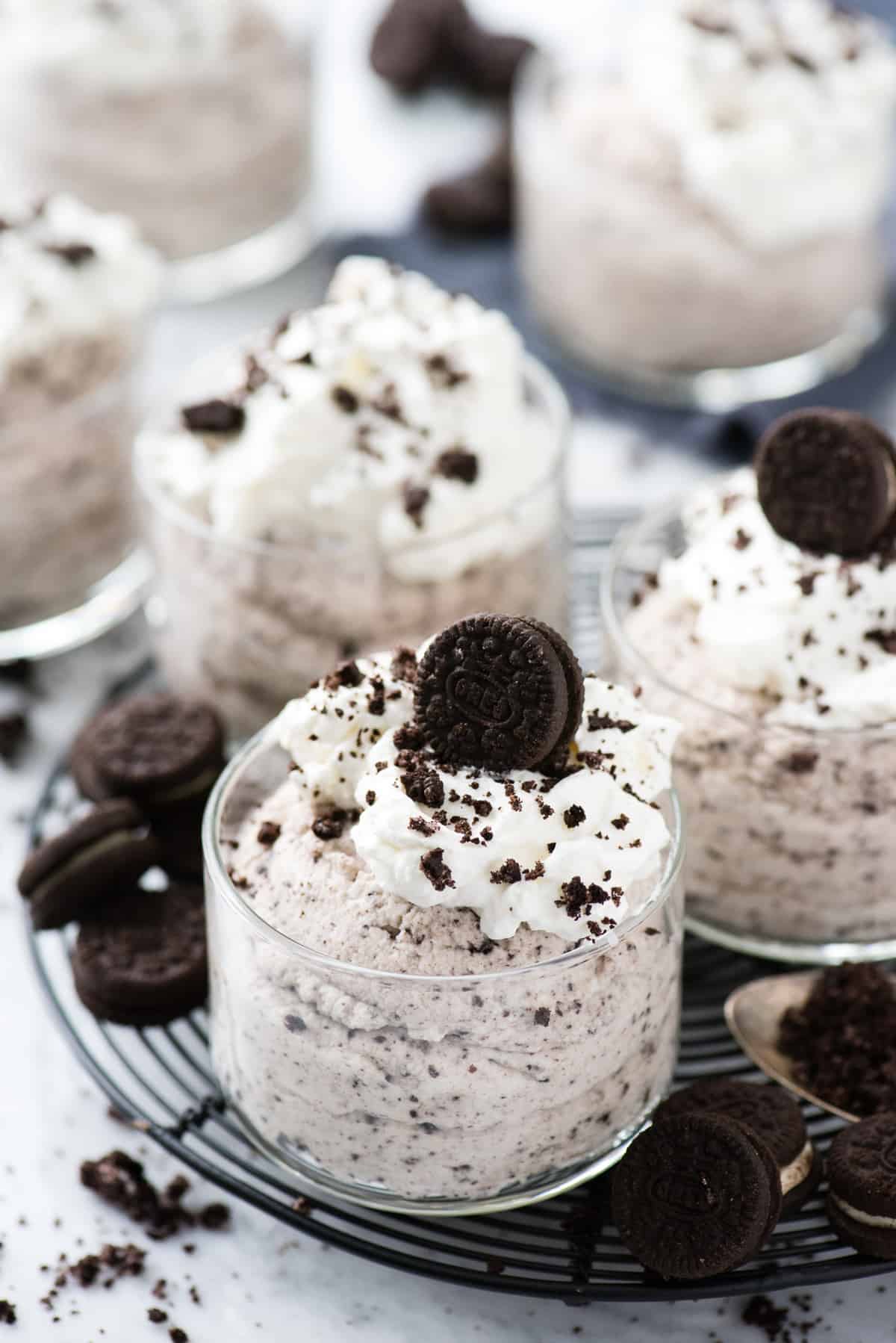 oreo mousse in glass dessert bowl topped with whipped cream and oreo pieces on black circle rack on white background