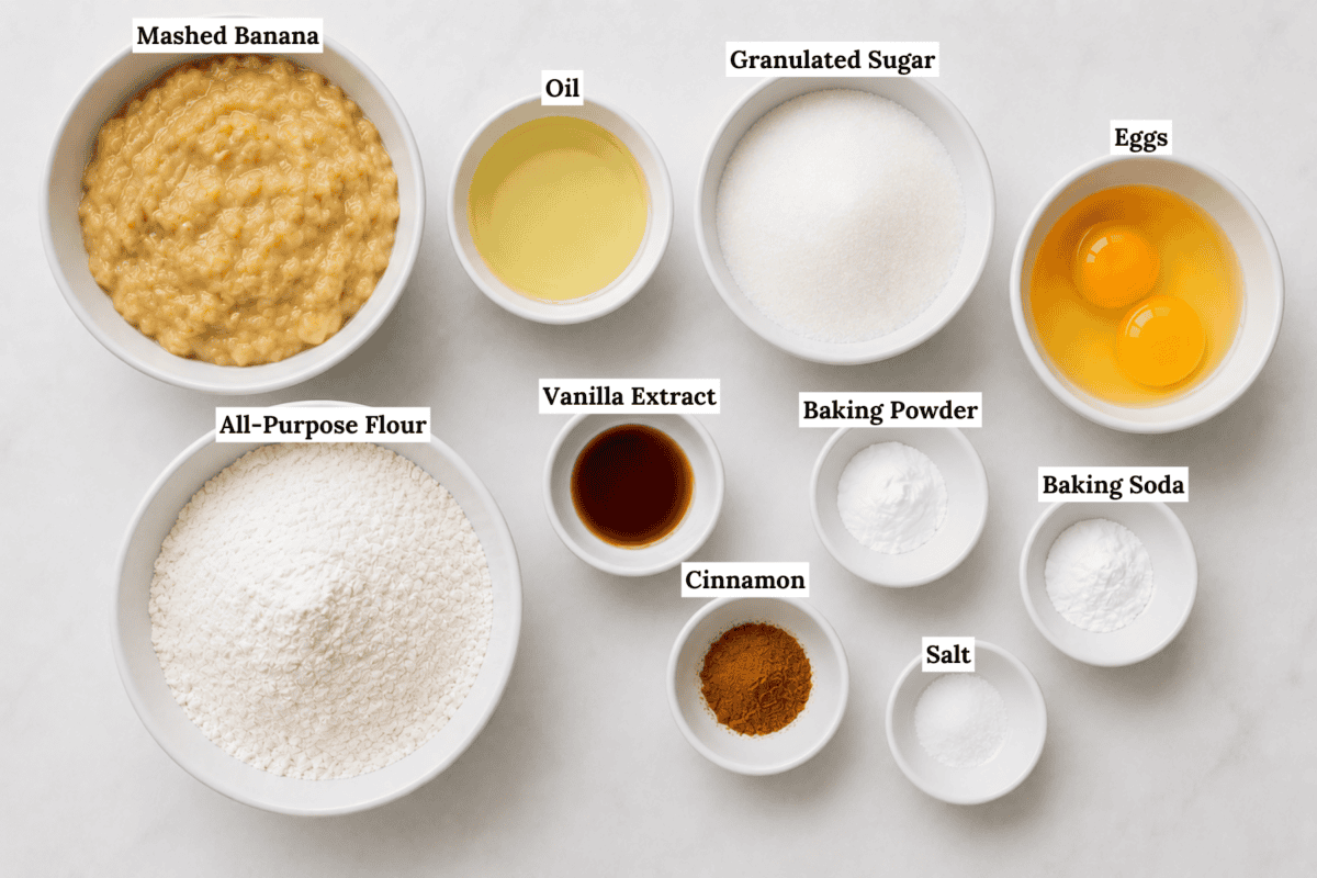 Bowls with labeled ingredients for easy banana bread: mashed banana, oil, granulated sugar, eggs, all-purpose flour, vanilla extract, baking powder, cinnamon, baking soda, and salt—neatly arranged on a white surface.