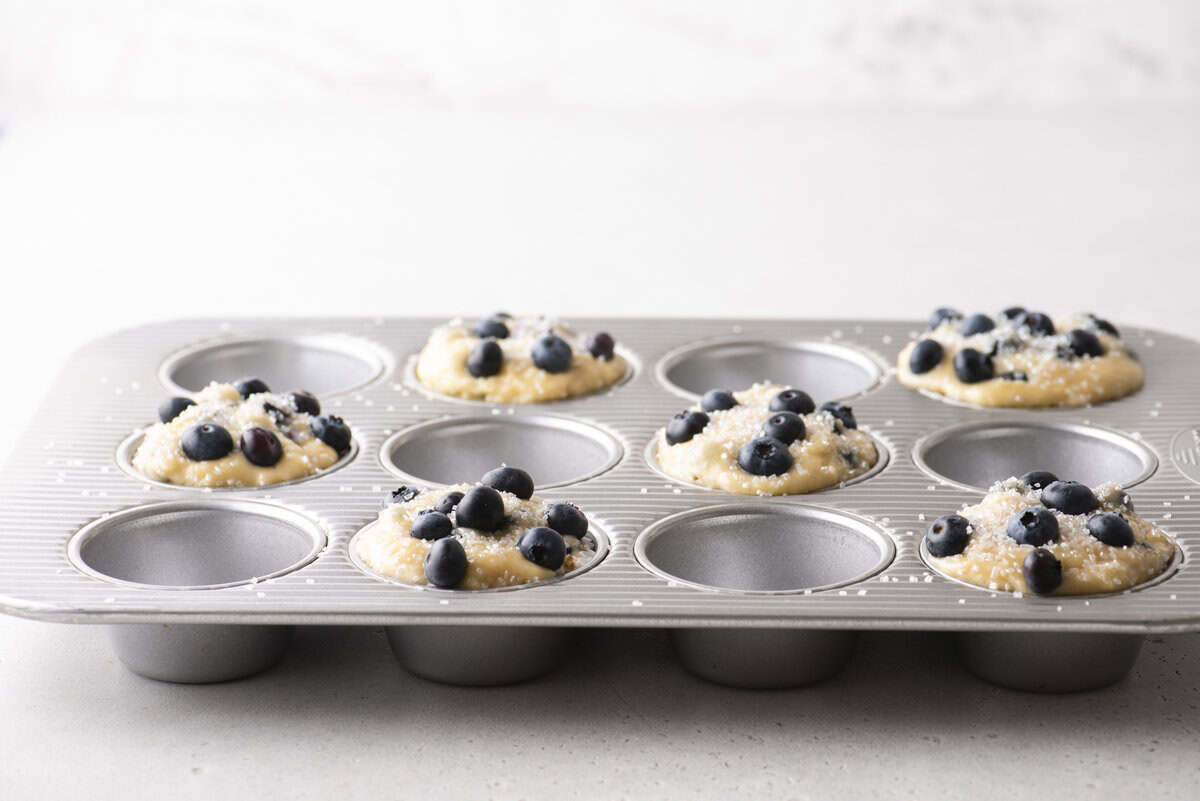 A metal muffin tray with nine cups, some filled with blueberry muffin batter and topped with fresh blueberries, sits on a light countertop, ready to be baked into delicious blueberry muffins.