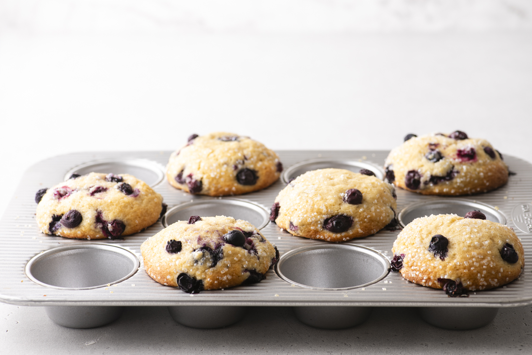 A muffin tin holds six golden blueberry muffins topped with coarse sugar. Two muffin spots are empty, showcasing the irresistible appeal of freshly baked blueberry muffins. The background is a light, neutral surface.