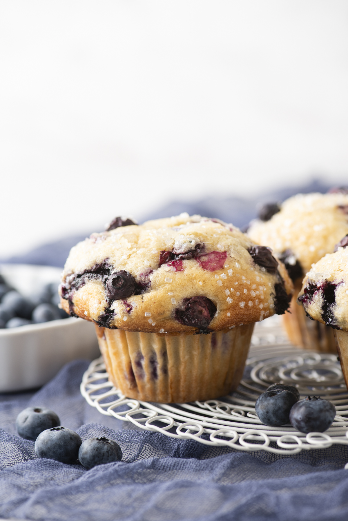 A close-up of a blueberry muffin topped with sugar crystals sits on a wire rack, highlighting the irresistible appeal of homemade blueberry muffins. Fresh blueberries and a bowl of berries are in the background on a blue cloth.