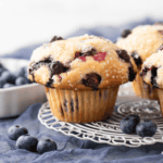 A close-up of freshly baked blueberry muffins with sugar crystals on top, sitting on a wire cooling rack with fresh blueberries and another muffin in the background.