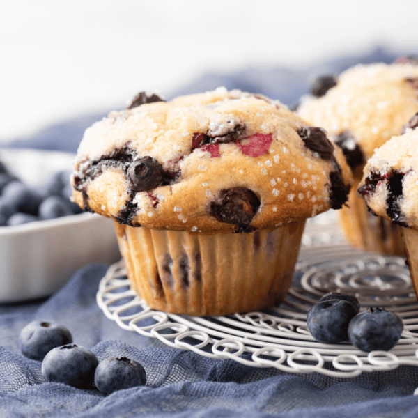 A close-up of freshly baked blueberry muffins with sugar crystals on top, sitting on a wire cooling rack with fresh blueberries and another muffin in the background.