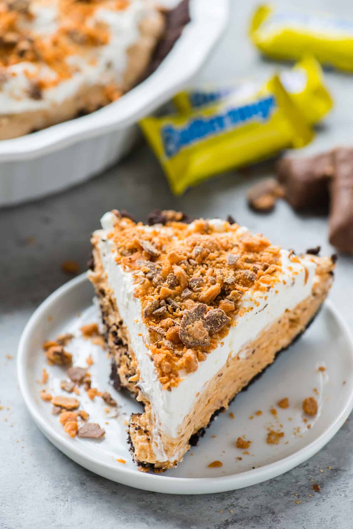 slice of butterfinger pie on white plate with pan of butterfinger pie in the background
