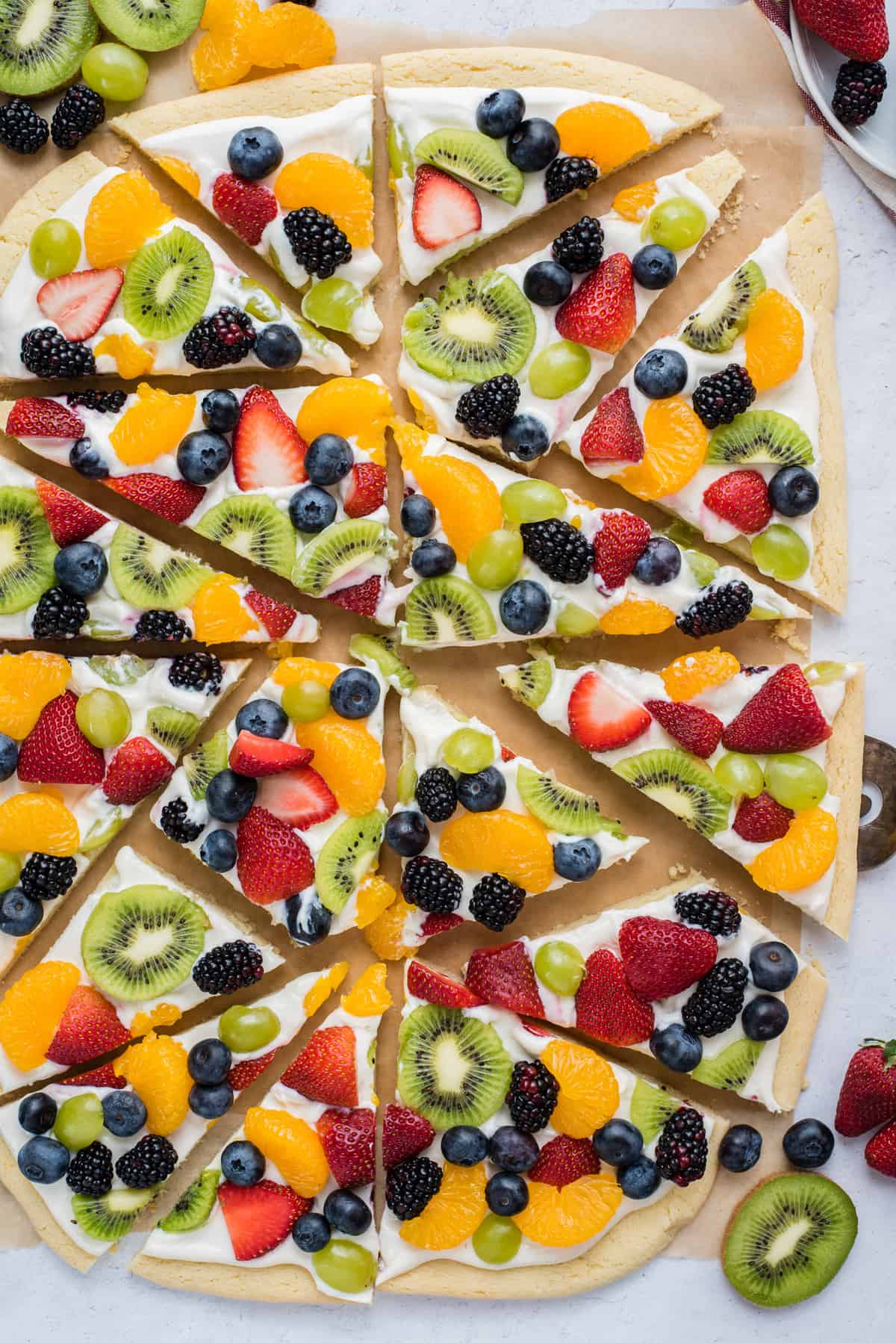 a whole fruit pizza cut into triangle slices with strawberries, blueberries, mandarin oranges, kiwis and grapes on brown parchment paper