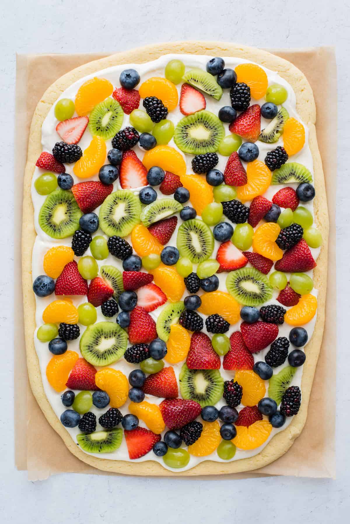 a whole fruit pizza with strawberries, blueberries, mandarin oranges, kiwis and grapes on brown parchment paper