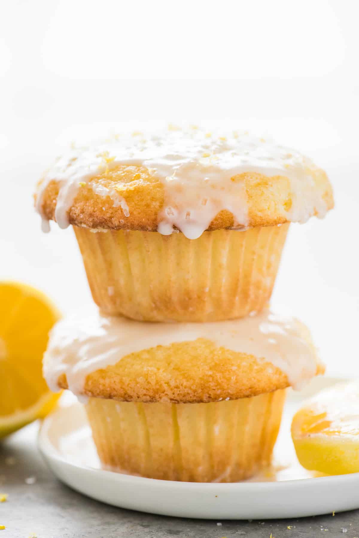 two lemon muffins with glaze stacked on each other on white plate on white background