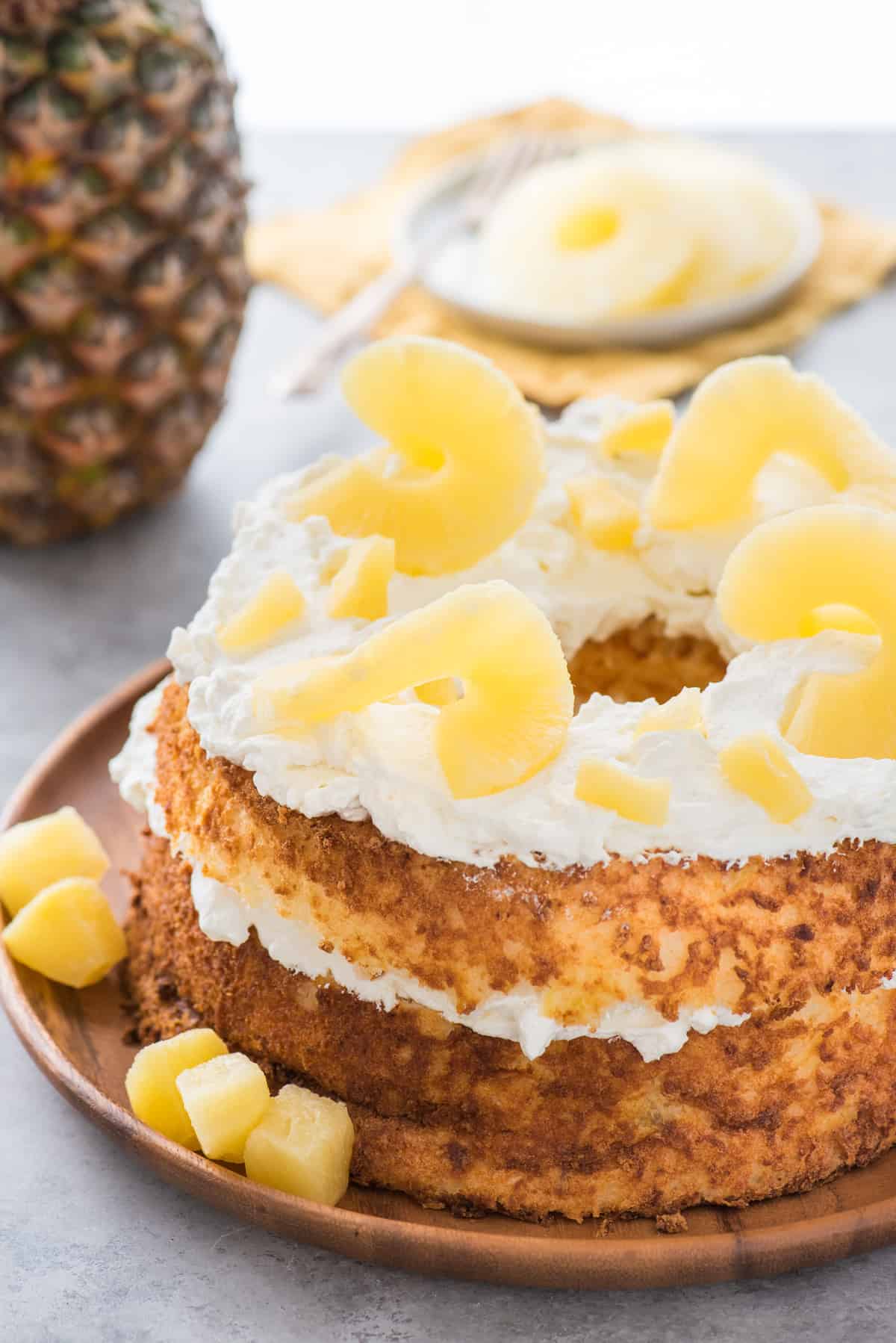 whole angel food cake topped with whipped cream and pineapple slices on wood serving tray on white background