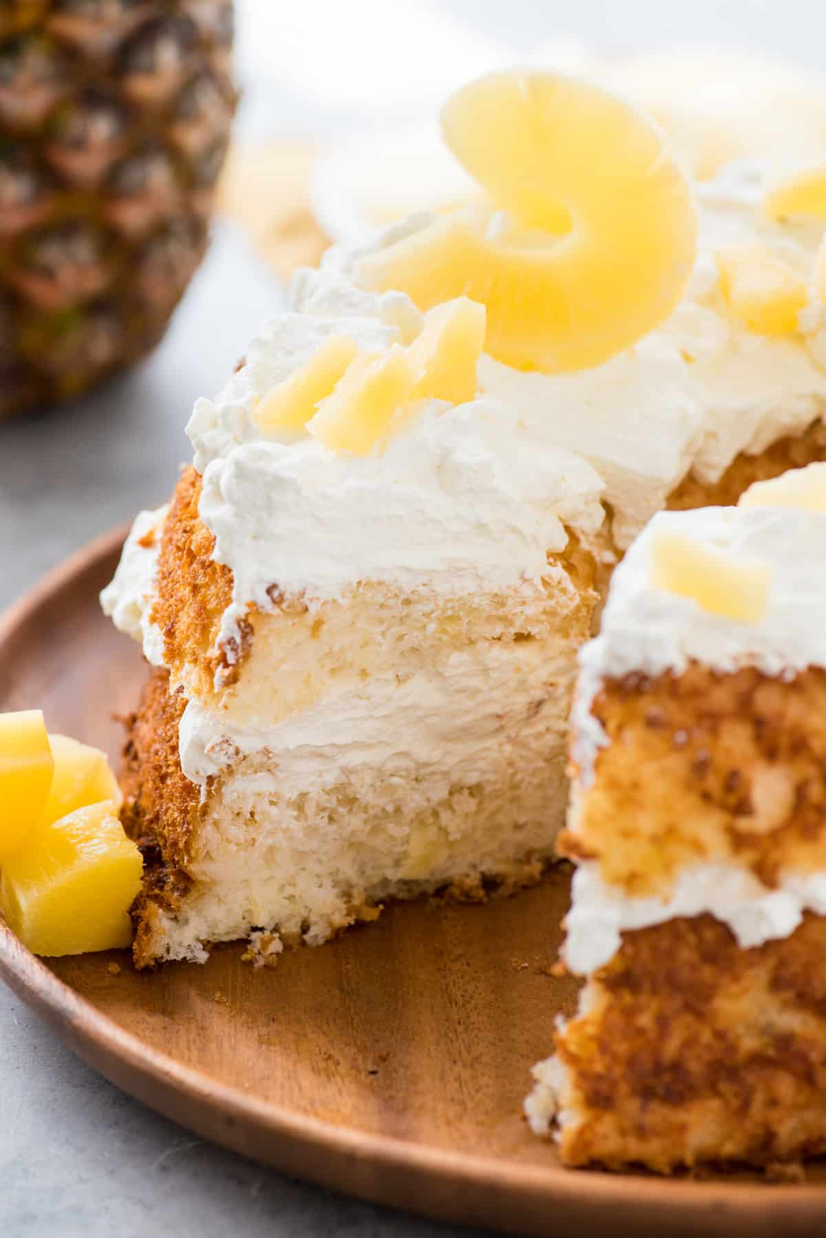 angel food cake with slice removed topped with whipped cream and pineapple slices on wood serving tray on white background