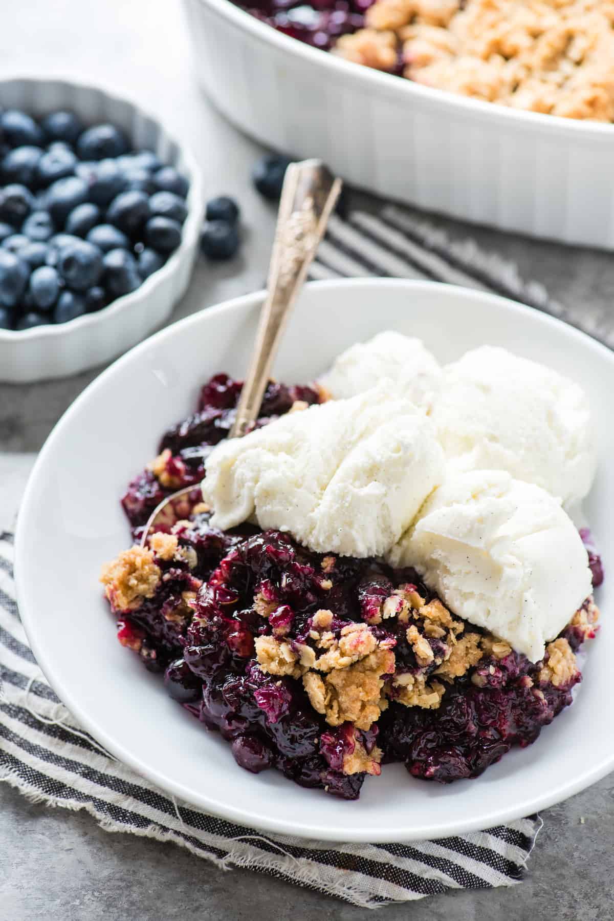 blueberry crisp in a white bowl with vanilla ice cream and a metal spoon with blue striped napkin underneath