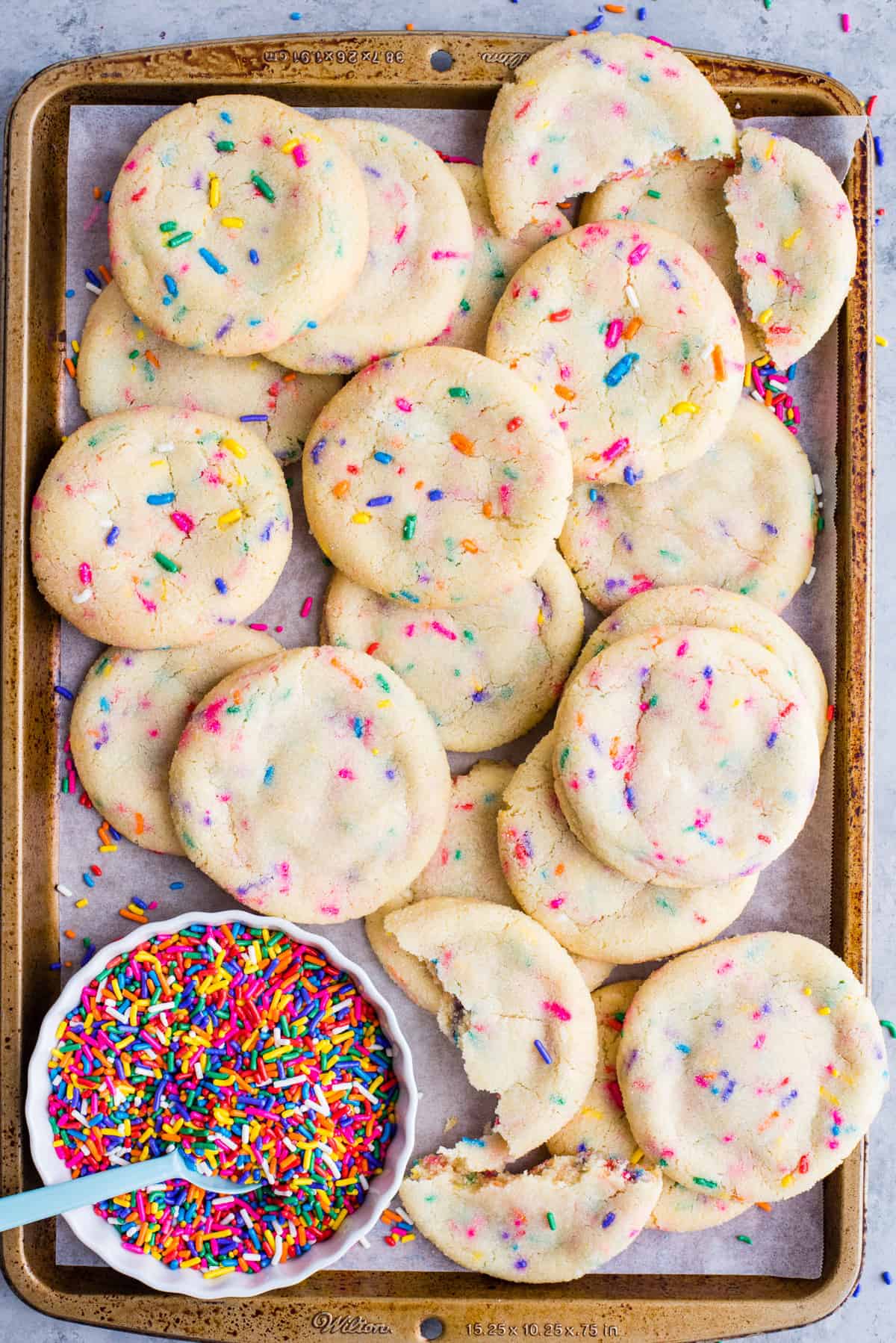 sprinkle sugar cookies arranged randomly on metal baking sheet with a bowl of rainbow sprinkles