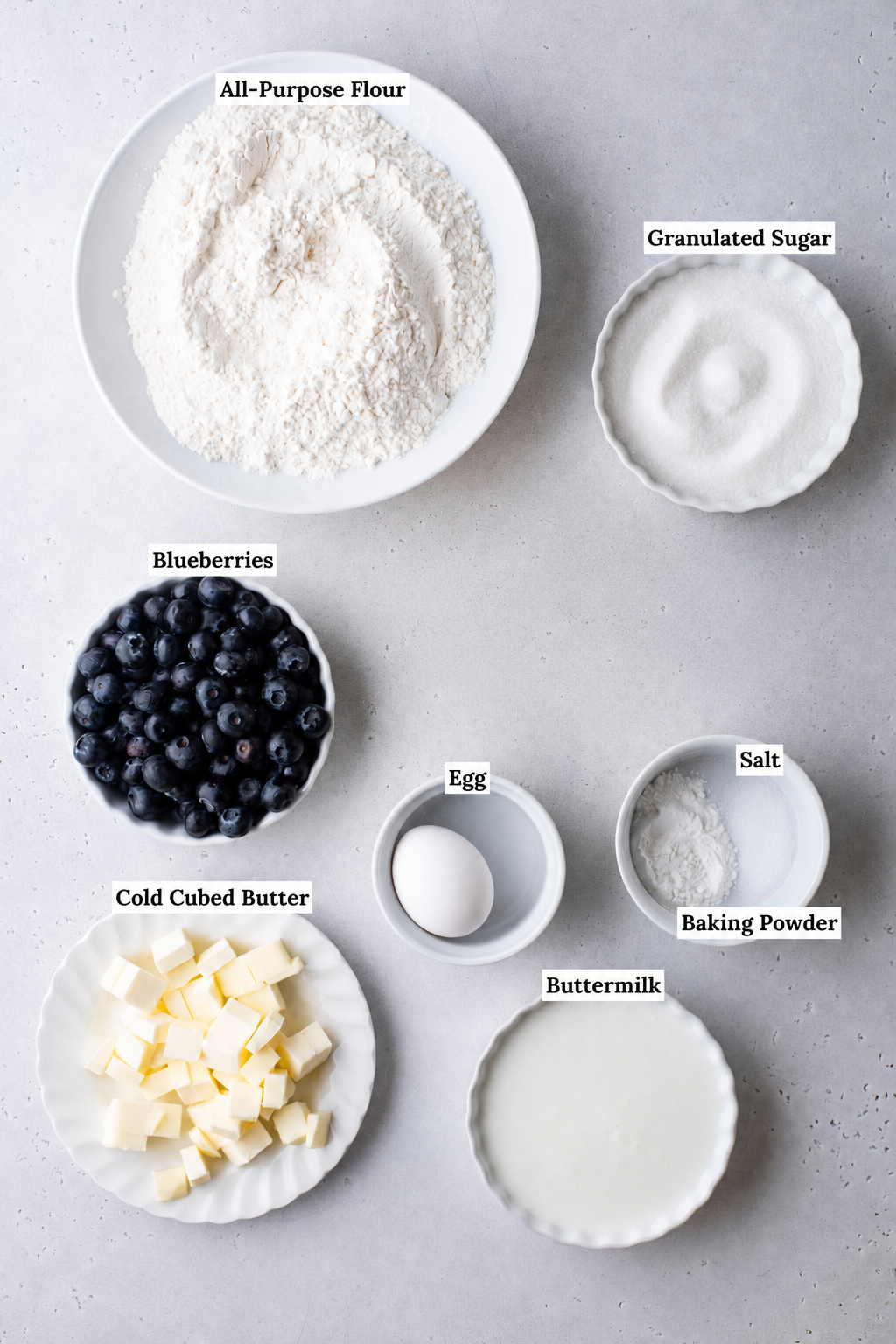 A flat lay of labeled ingredients in bowls for baking blueberry scones: all-purpose flour, granulated sugar, blueberries, a whole egg, cubed cold butter, buttermilk, salt, and baking powder.