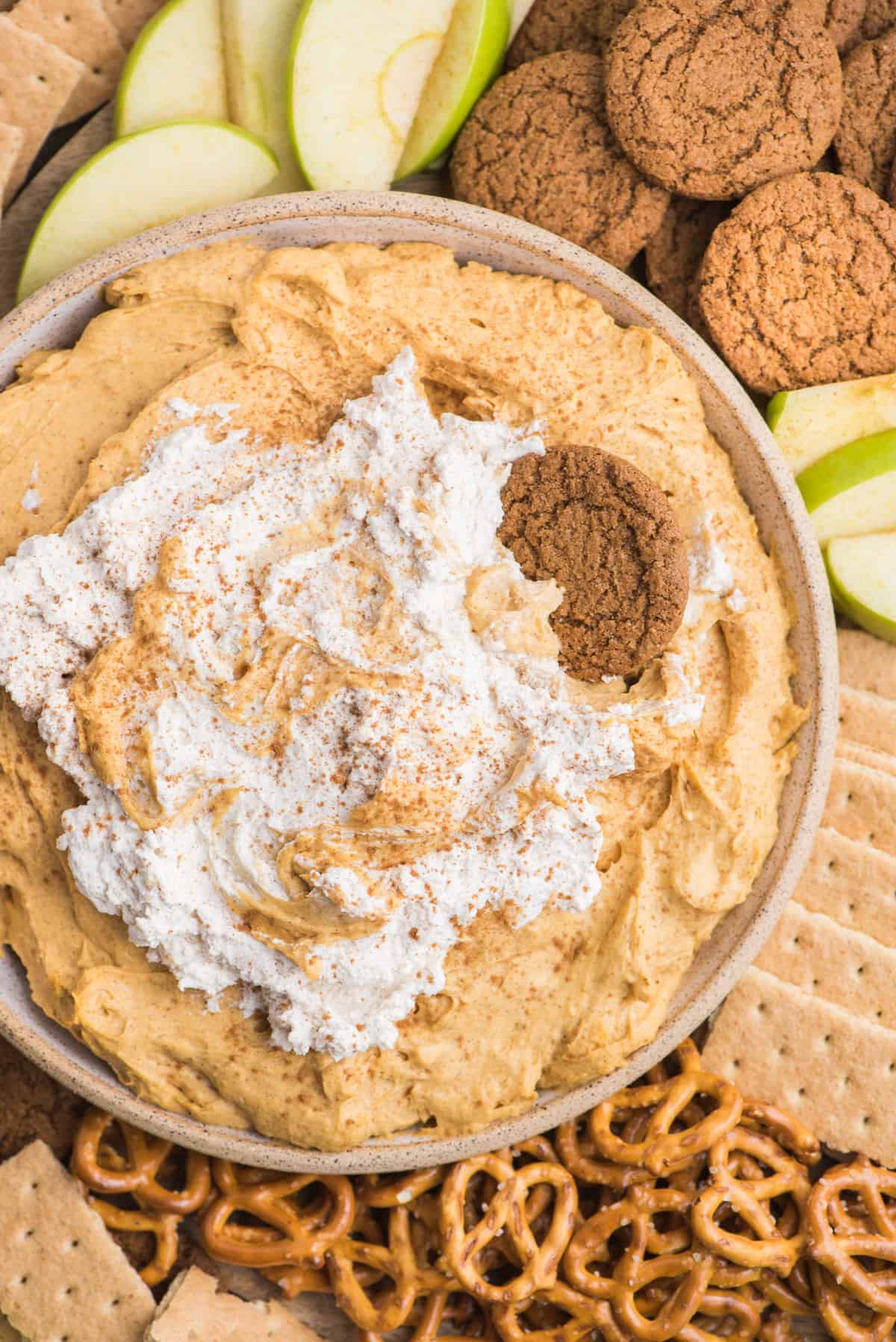 pumpkin dip with whipped cream topping in a bowl surrounded by piles of pretzels, gingersnaps, graham crackers and green apple slices