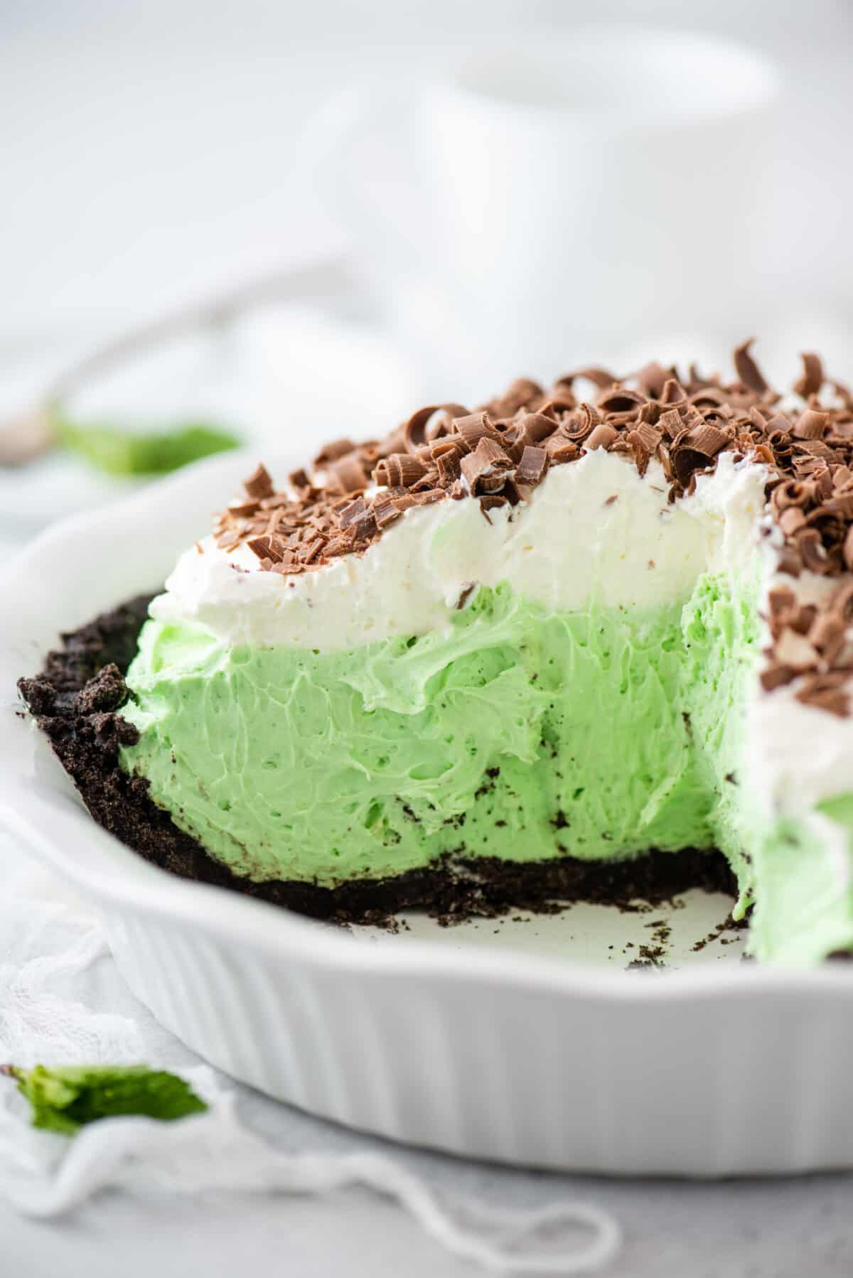A close-up of a chocolate mint pie in a white dish, featuring a green mint filling on an Oreo pie crust, topped with whipped cream and chocolate shavings, with a slice removed.