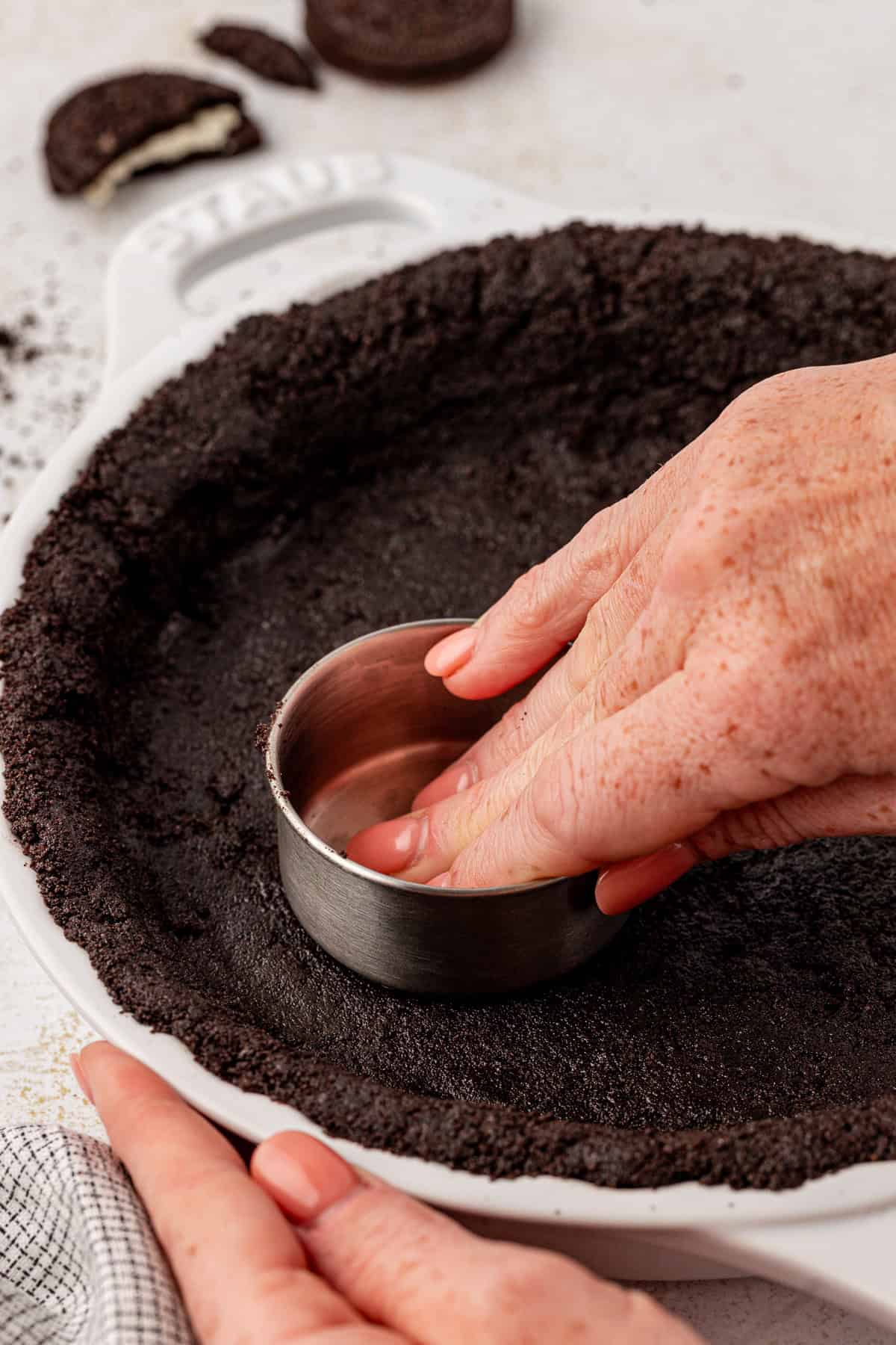 Close-up of hands pressing a metal measuring cup into a pie dish to form an Oreo pie crust from crushed chocolate cookies. Broken cookies are visible in the background.