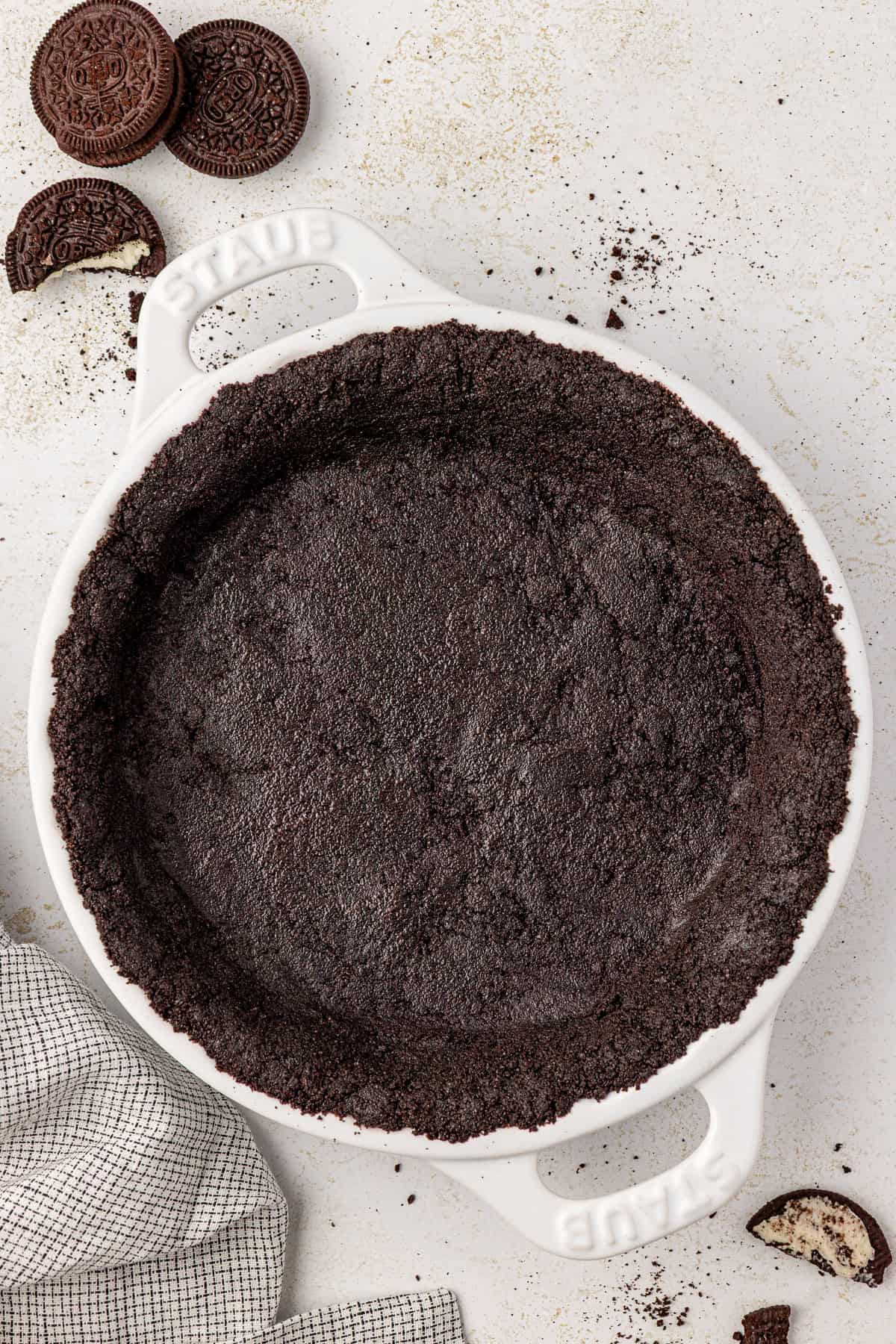A white pie dish filled with a pressed Oreo pie crust sits on a light surface. Crushed cookie crumbs are scattered nearby, with whole and broken Oreo cookies and a folded dish towel beside the dish.