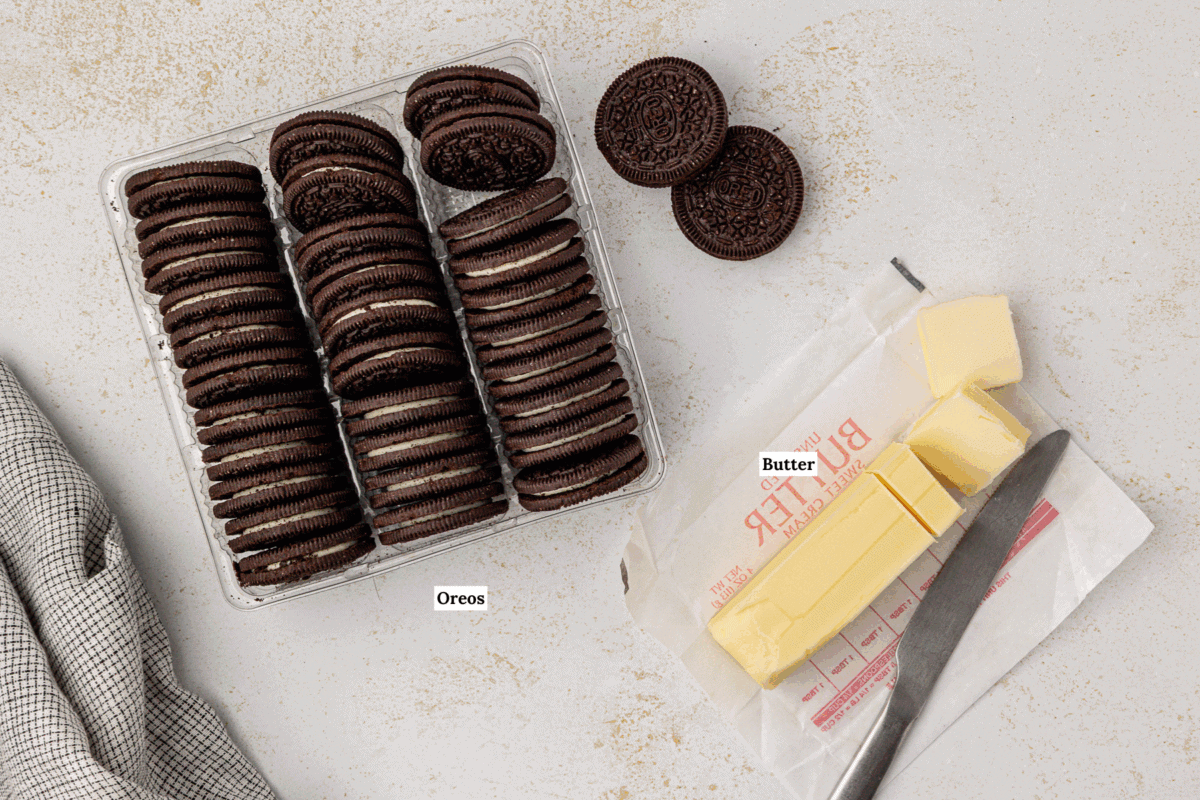 A package of chocolate sandwich cookies and a stick of butter, labeled “Oreos” and “Butter,” sit on a light countertop—perfect for making an Oreo pie crust. A gray dish towel is also visible on the side.