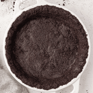 A white pie dish filled with a pressed Oreo pie crust is shown from above. The crust edges are evenly shaped, and the dish sits on a light surface with part of a checkered towel visible nearby.