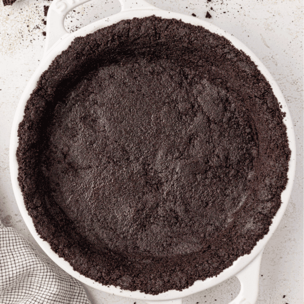 A white pie dish filled with a pressed Oreo pie crust is shown from above. The crust edges are evenly shaped, and the dish sits on a light surface with part of a checkered towel visible nearby.
