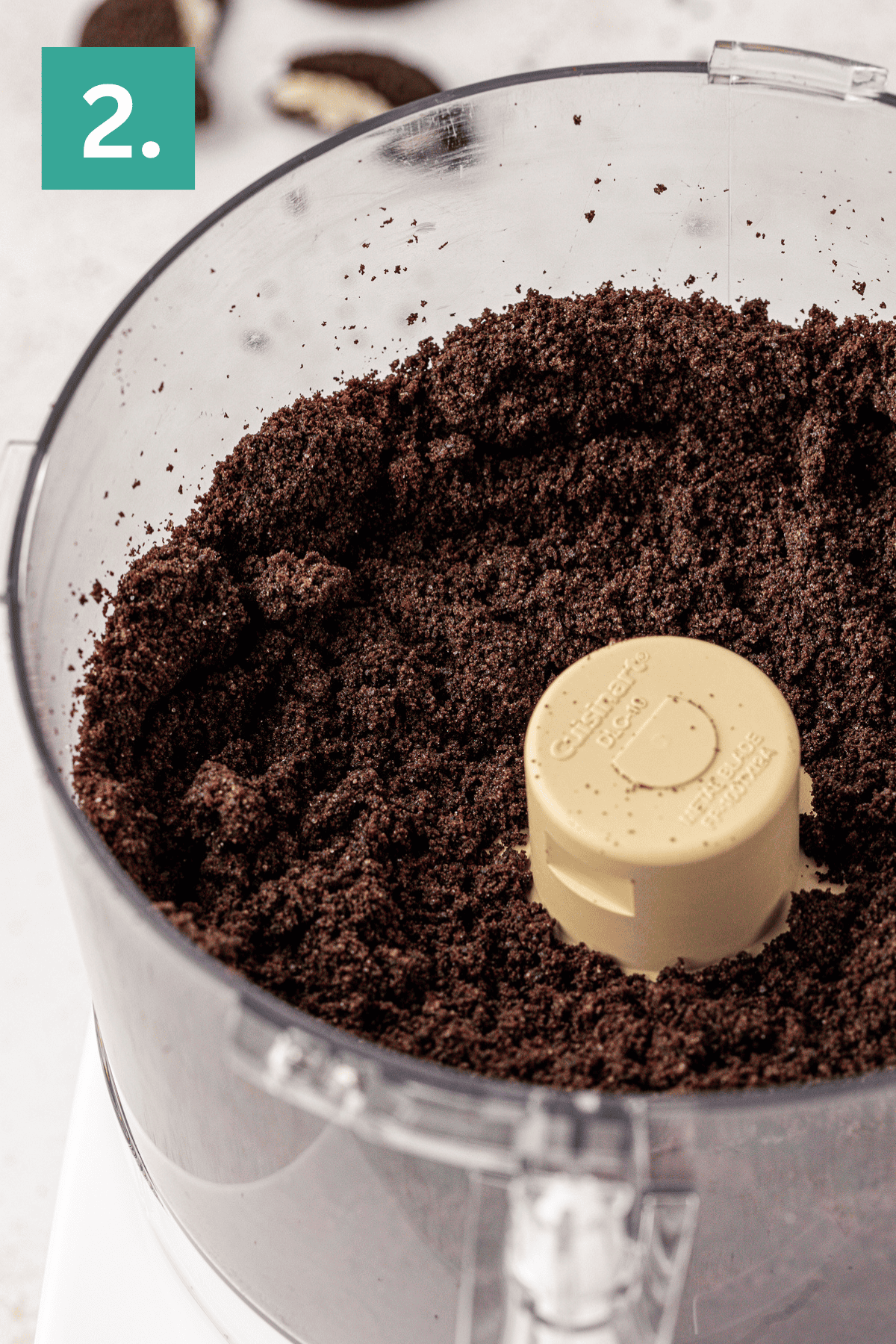 Close-up of a food processor filled with finely crushed chocolate cookies for an Oreo pie crust, shown as step 2 in a recipe. A teal box with the number 2 appears in the upper left corner.